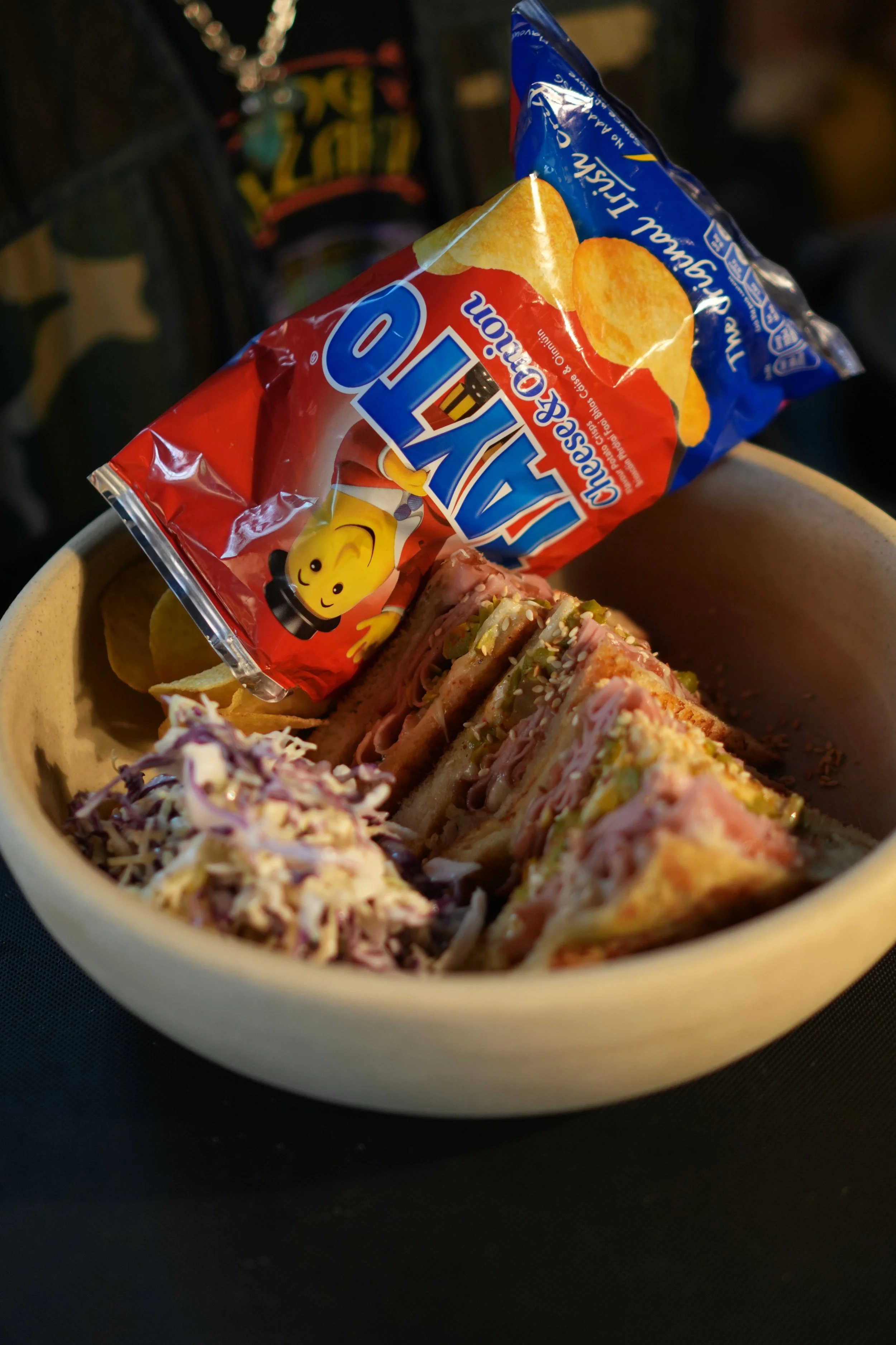A bowl with sandwiches topped with various ingredients, including slaw and sesame seeds, and a bag of Tayto's potato chips.