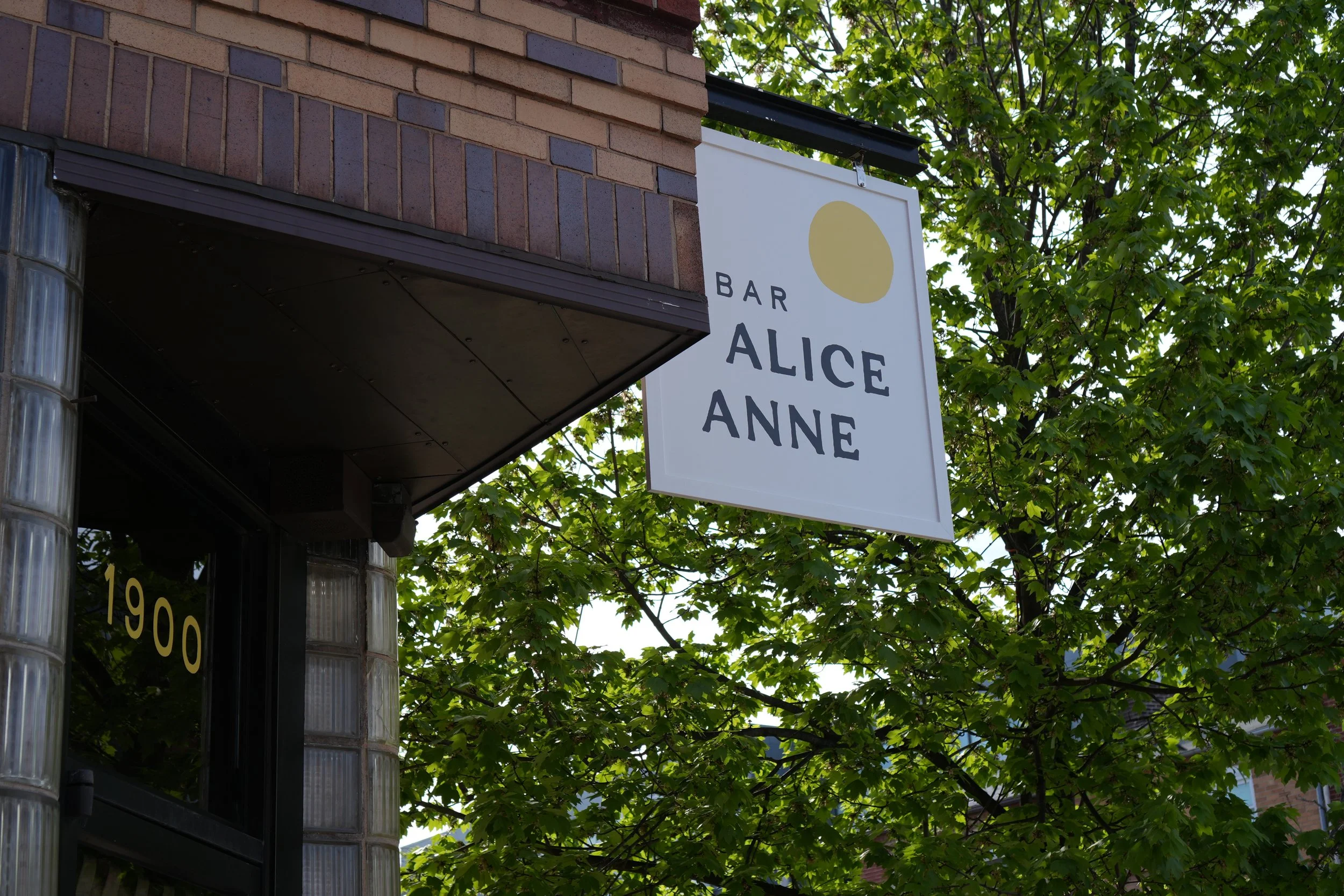 Sign for Bar Alice Anne hanging outside a building with brick and glass siding, surrounded by green leafy trees