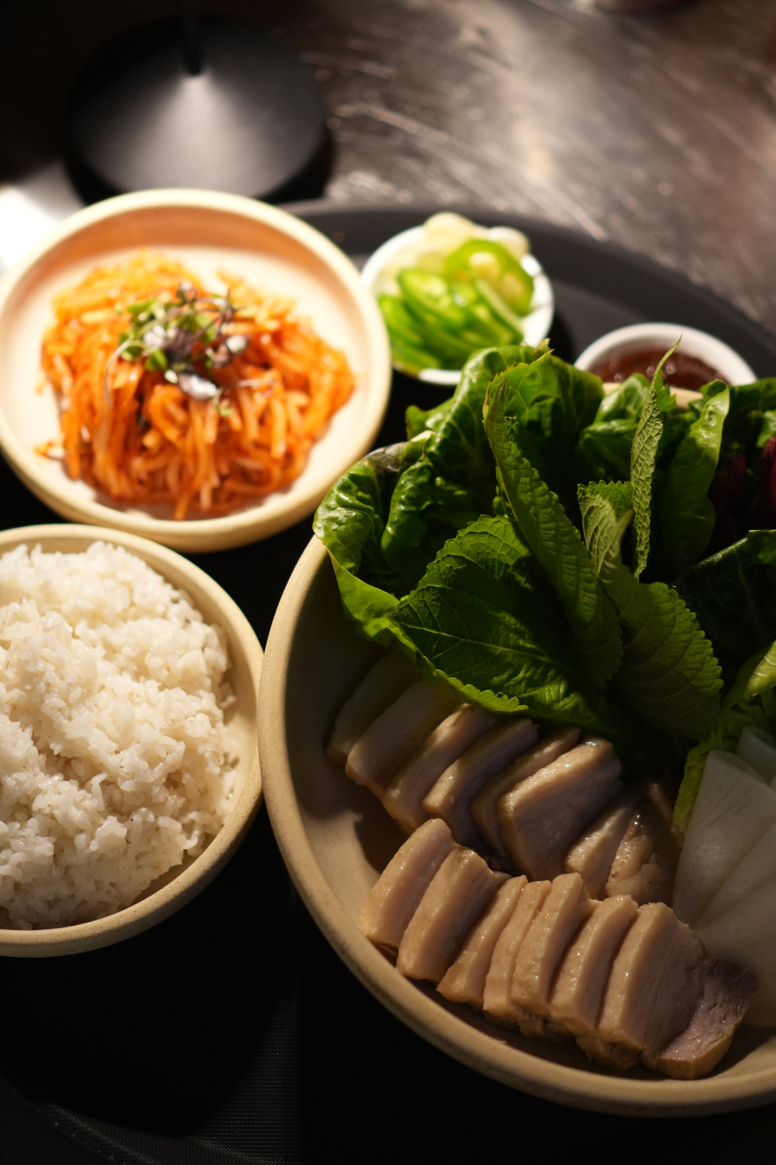 A Korean ssam plate meal with rice, sliced boiled pork belly, fresh green lettuce, cucumber salad, kimchi, and dipping sauces.
