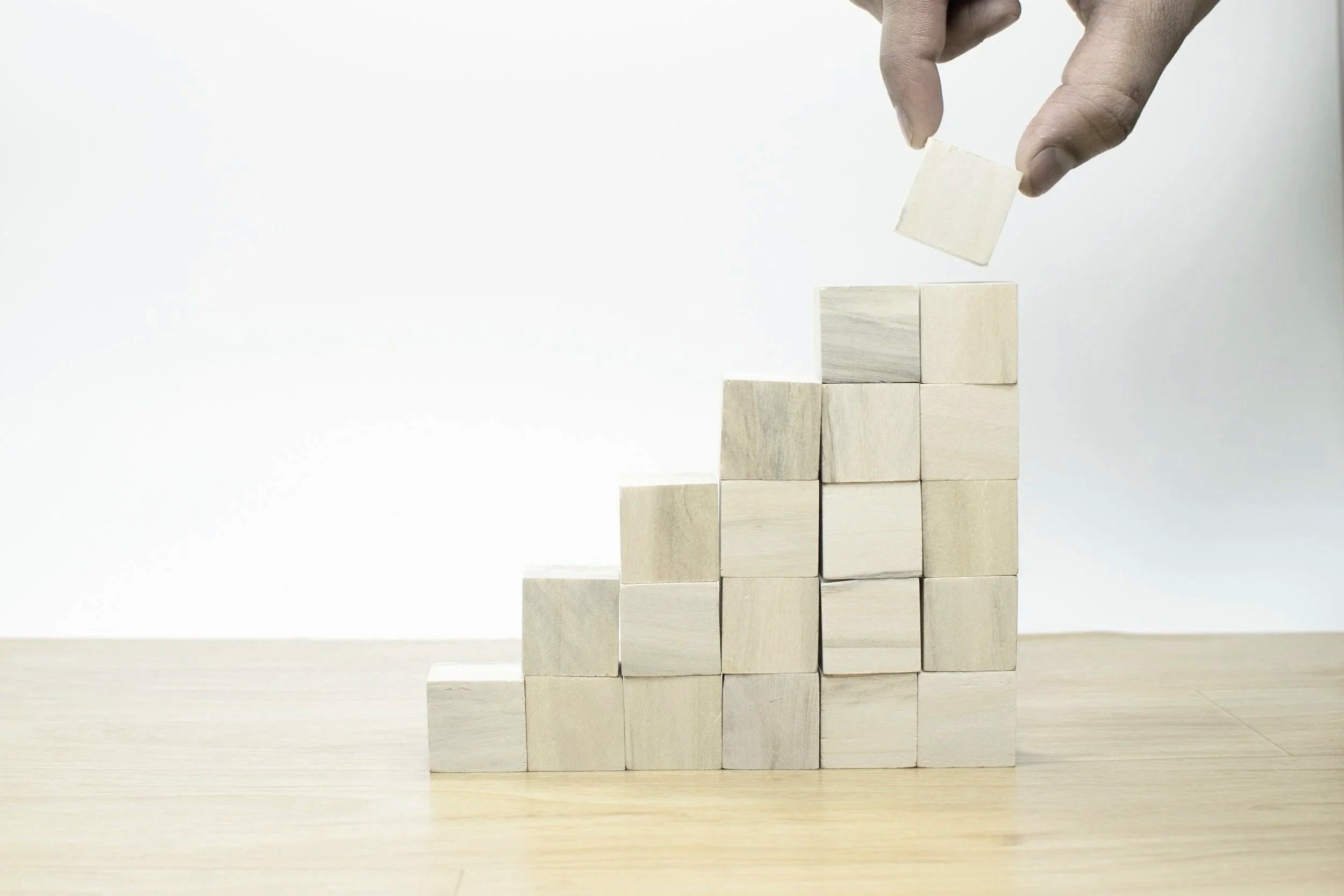 A hand placing a wooden cube on top of a stack of similar cubes, forming a small pyramid against a plain background.