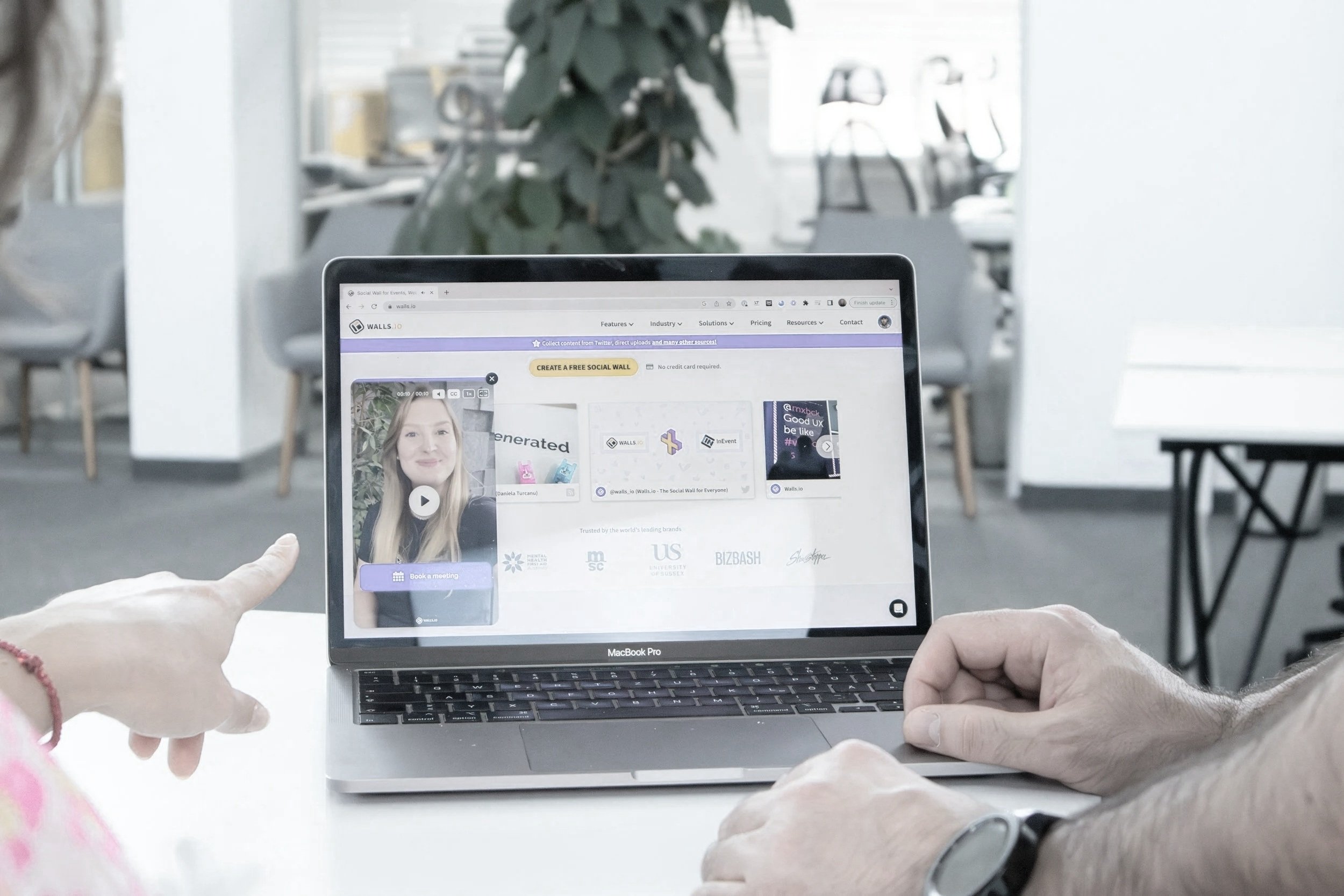 Person holding a MacBook Pro displaying a website for creating social media walls, with another person pointing at the screen in a bright office space.