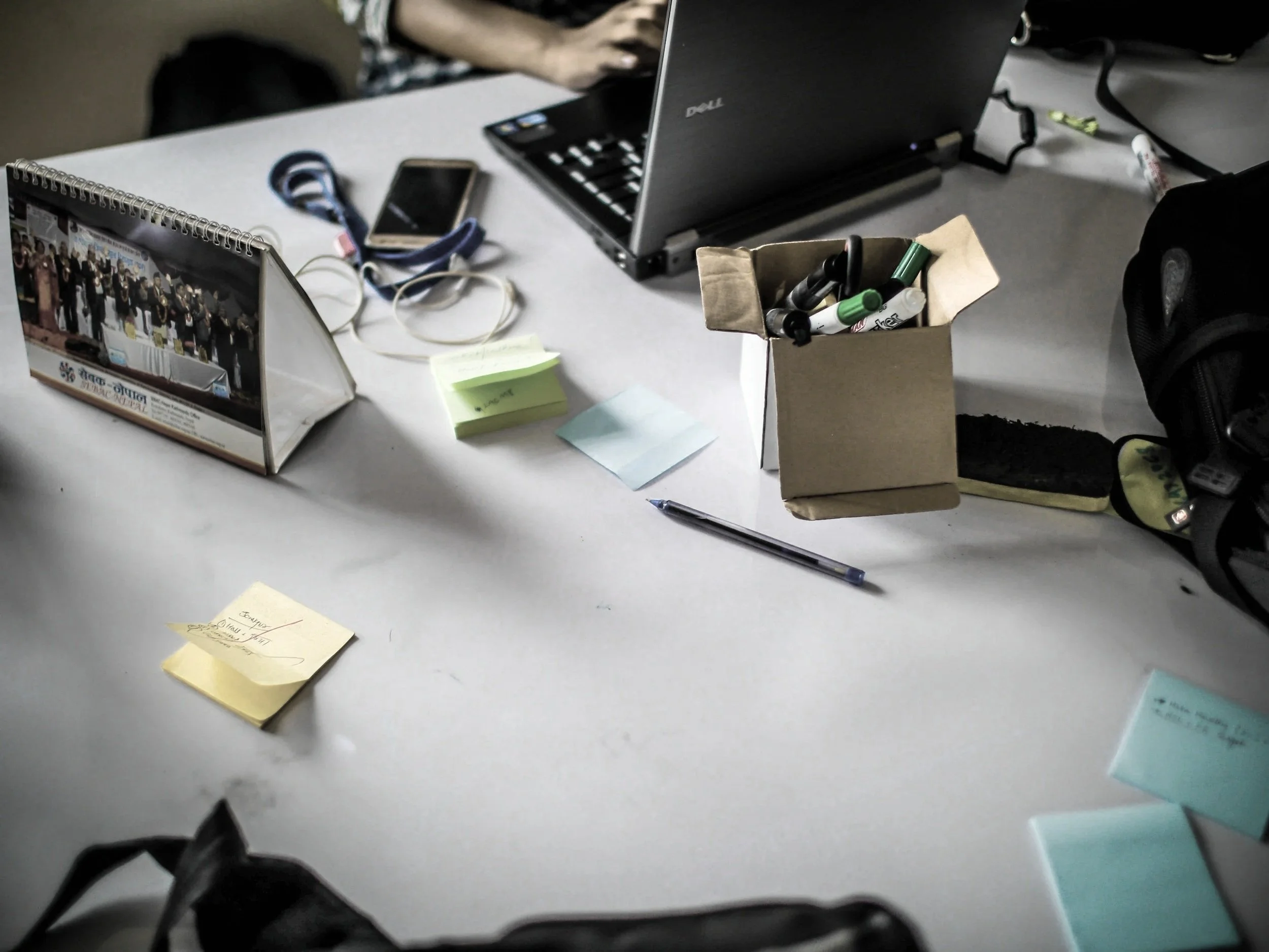 A cluttered office desk with a laptop, a mobile phone, a pen, sticky notes, markers in a small cardboard box, a calendar, and bags or backpacks in the background.