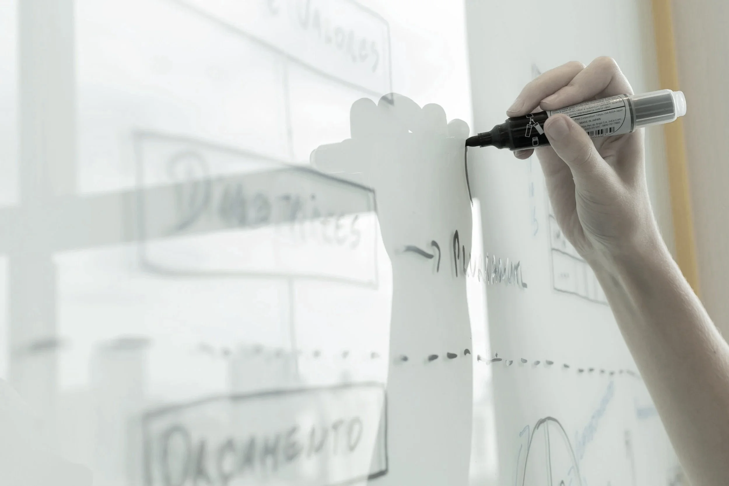 A person wearing gloves is writing with a black marker on a transparent whiteboard, which has some text and diagrams drawn on it.