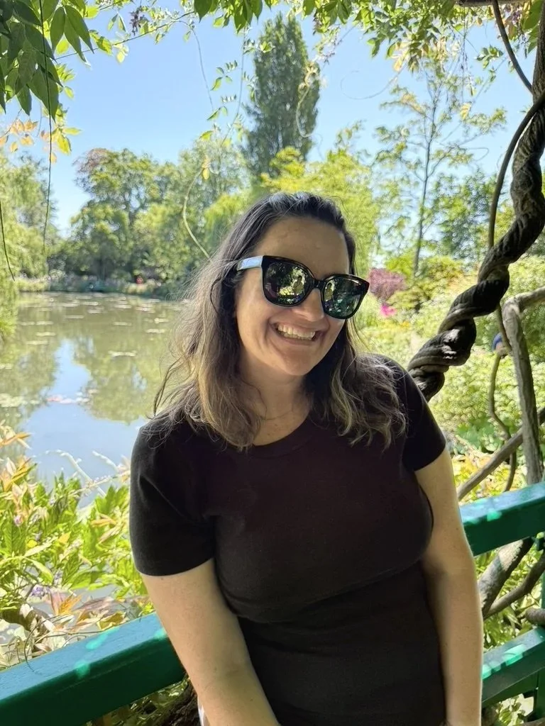 A woman with dark hair, wearing sunglasses and a black t-shirt, smiling outdoors near a river or pond with trees and greenery in the background.