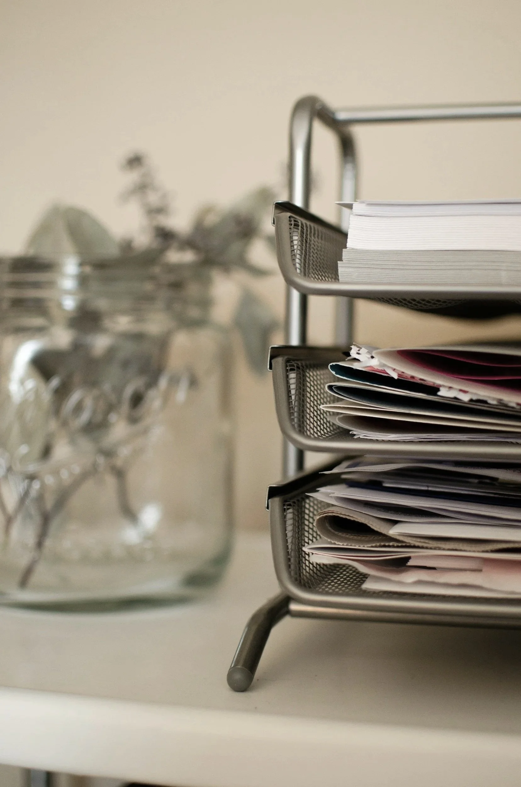 A desk with a metal organizer filled with papers and folders. Behind it, a blurred glass container holding stainless steel spoons and a plate in the background.