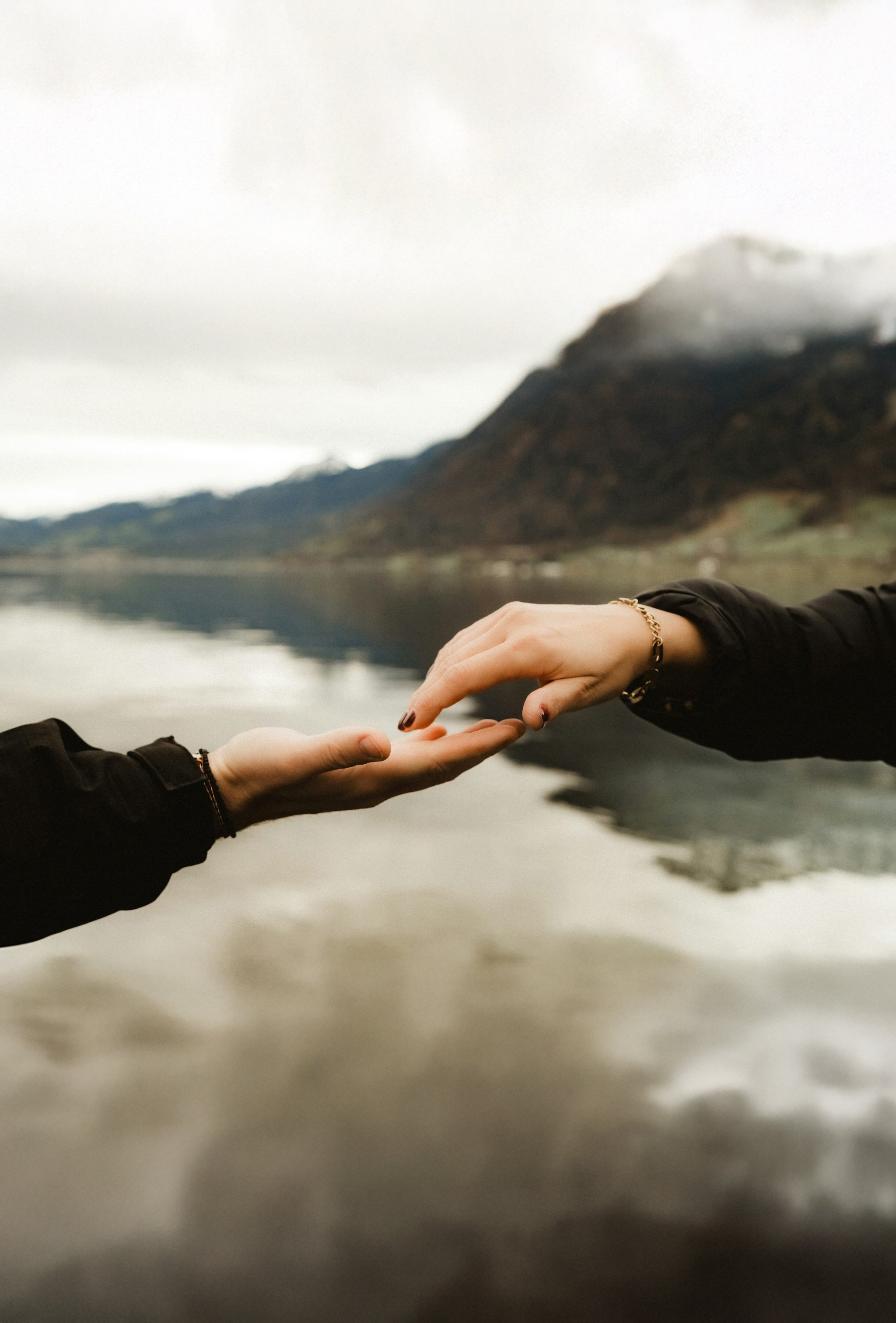 Two hands reaching towards each other over a reflective body of water with a mountain and cloudy sky in the background.