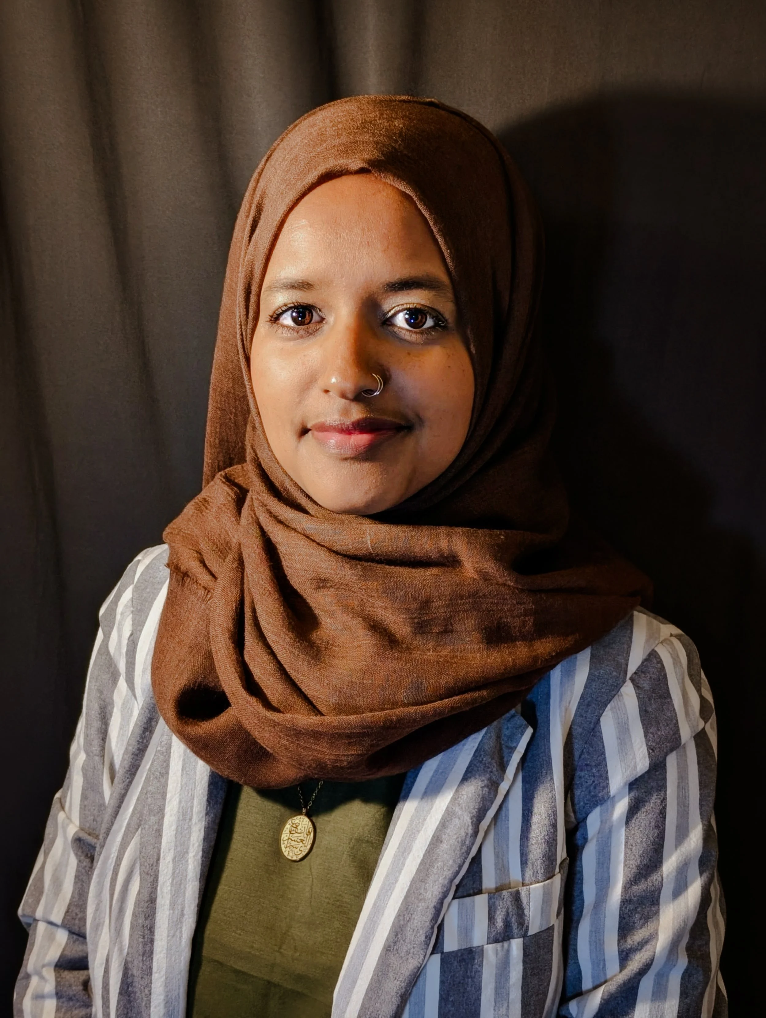 Portrait of a woman wearing a brown hijab, striped blazer, and green top, with a gold pendant necklace, standing against a dark background.