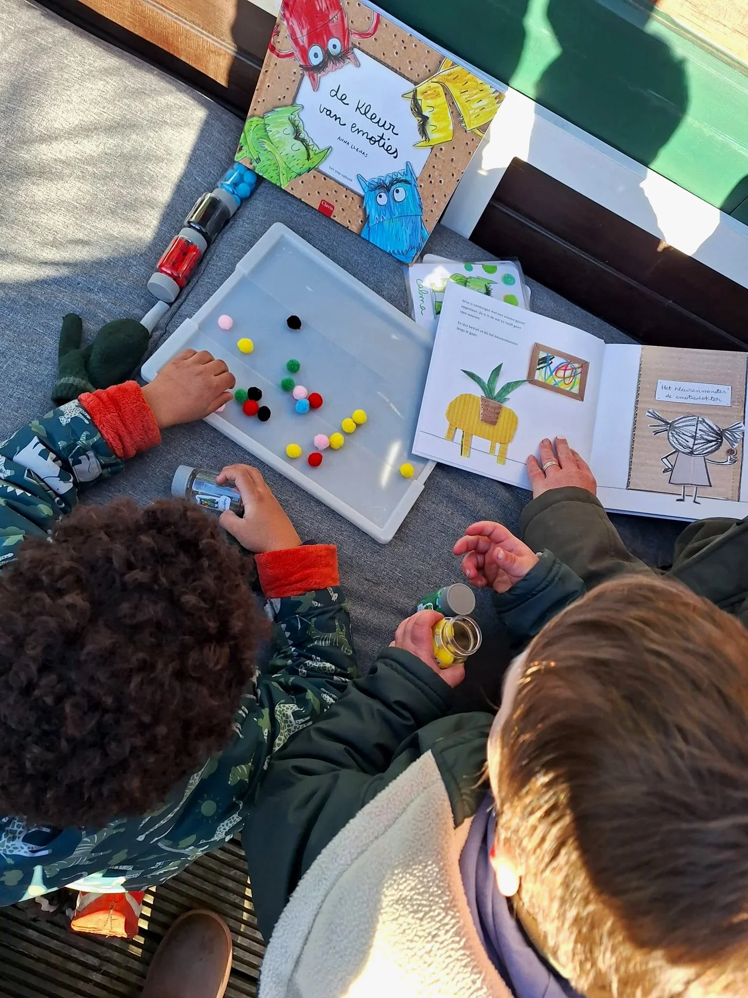 Twee kinderen die knutselactiviteiten doen met een boek over kleuren en een tray met kleurrijke pompoms en verf. Een boek met tekeningen en tekst ligt open.