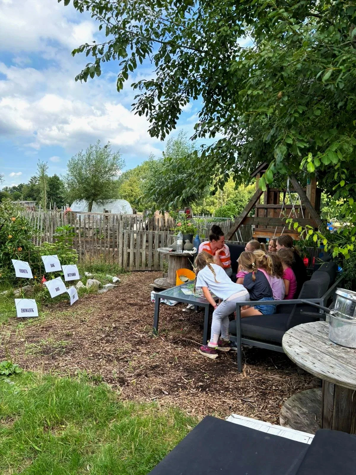 Gezelschap van kinderen en een volwassene buiten onder een boom, zittend op banken en aan tafel, met een tuin en houten schutting op de achtergrond.