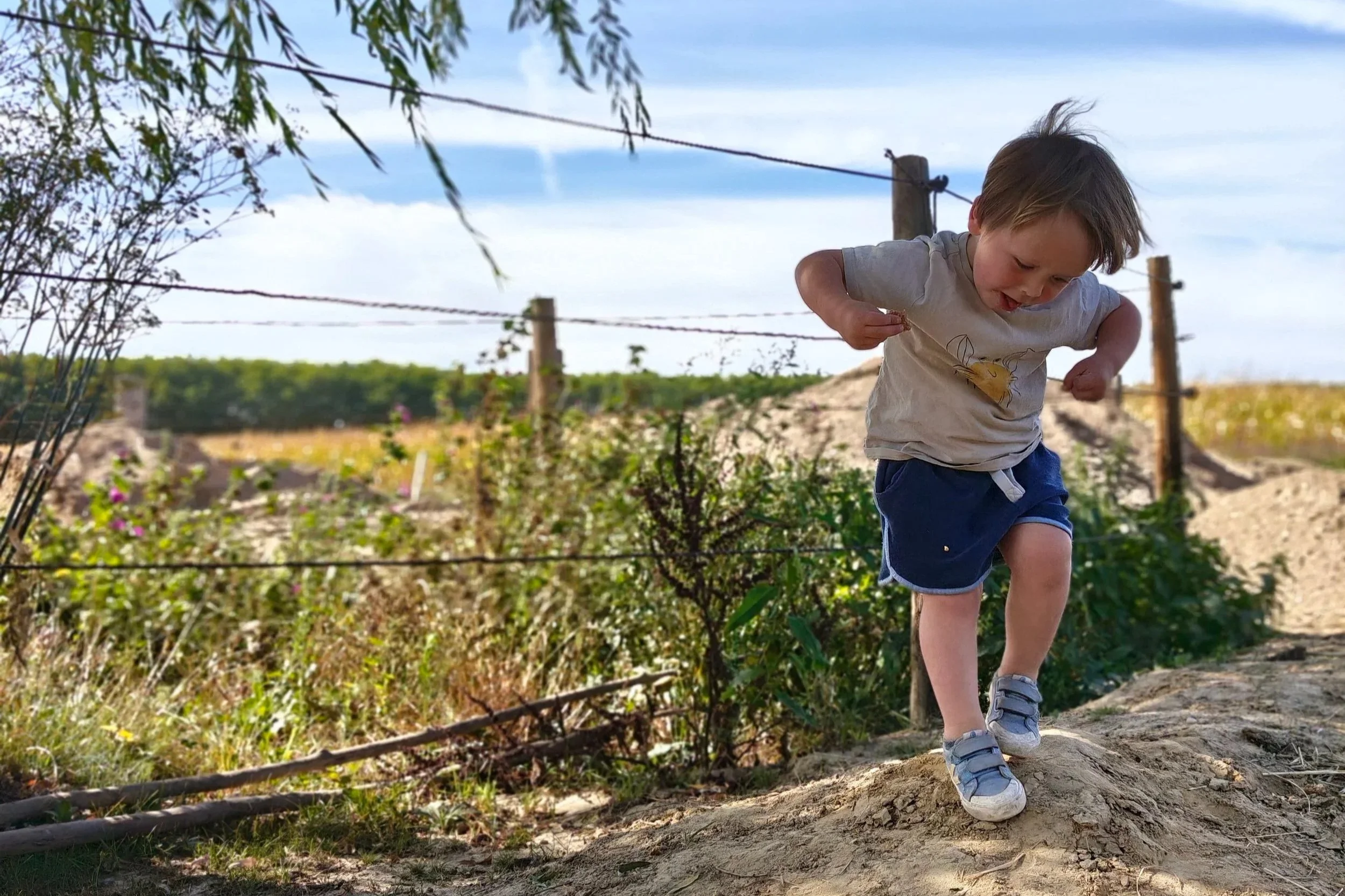 Een jonge jongen rent en huppelt op een zandige steile helling buiten, met een boomgaard of akker op de achtergrond, onder een heldere blauwe lucht.