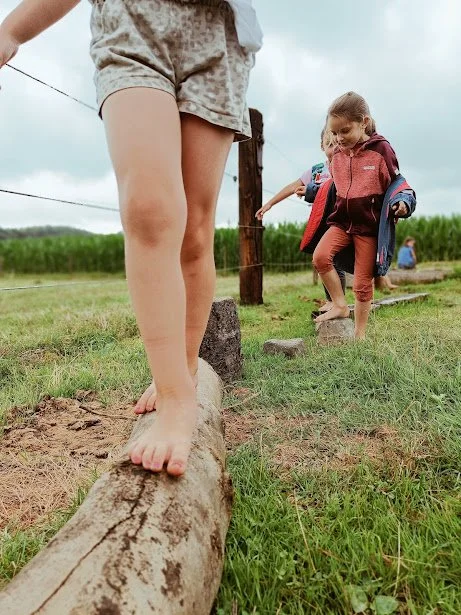 Kinderen lopen over een houten balk in een grasveld, met een meisje die een rode jas draagt en een jongen die achter haar loopt.