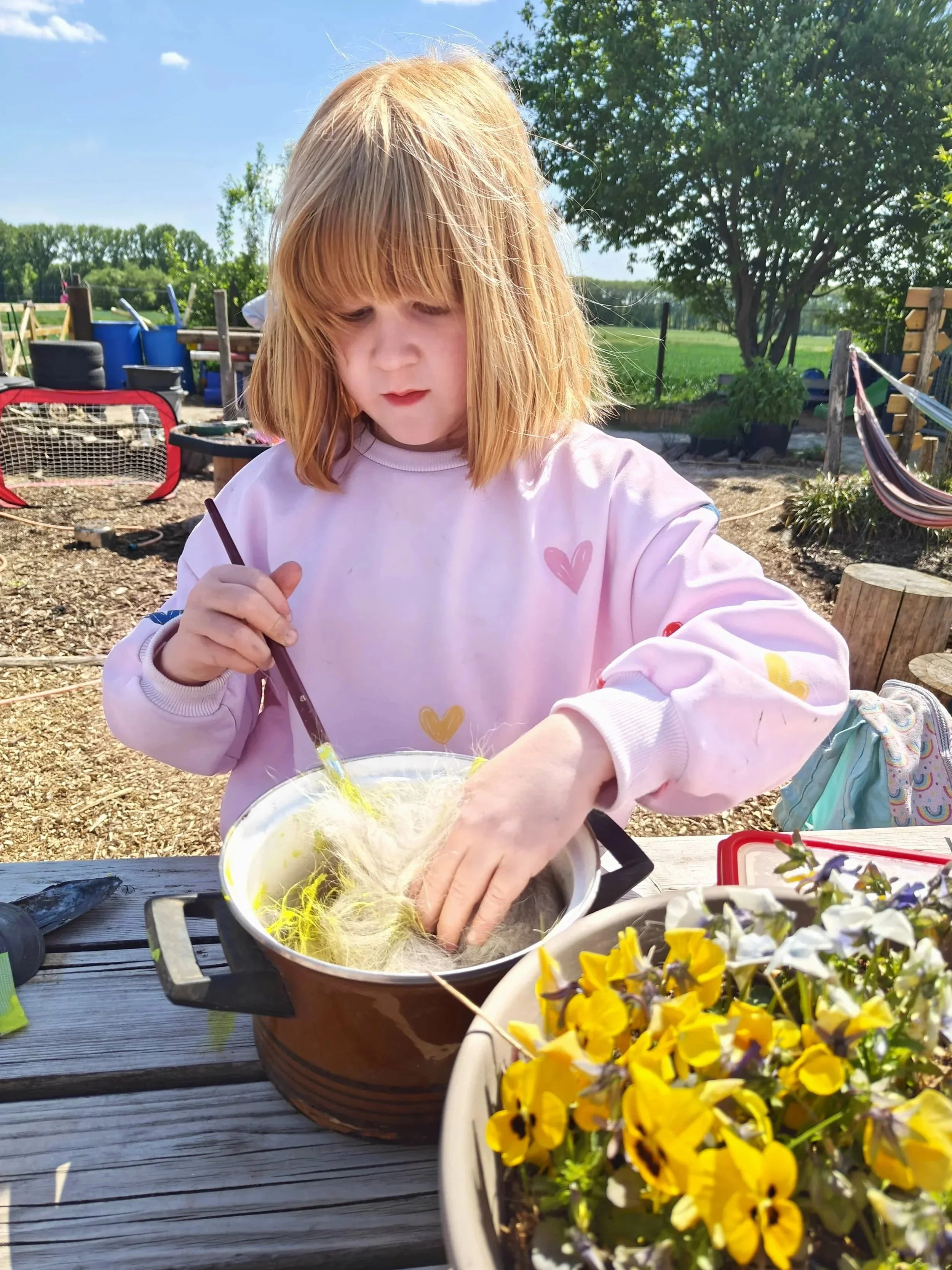 Een meisje met rood haar en een roze sweatshirt onderzoekt een pot met spunvlies of fiber, buiten op een houten tafel, met bloemen en tuin op de achtergrond.
