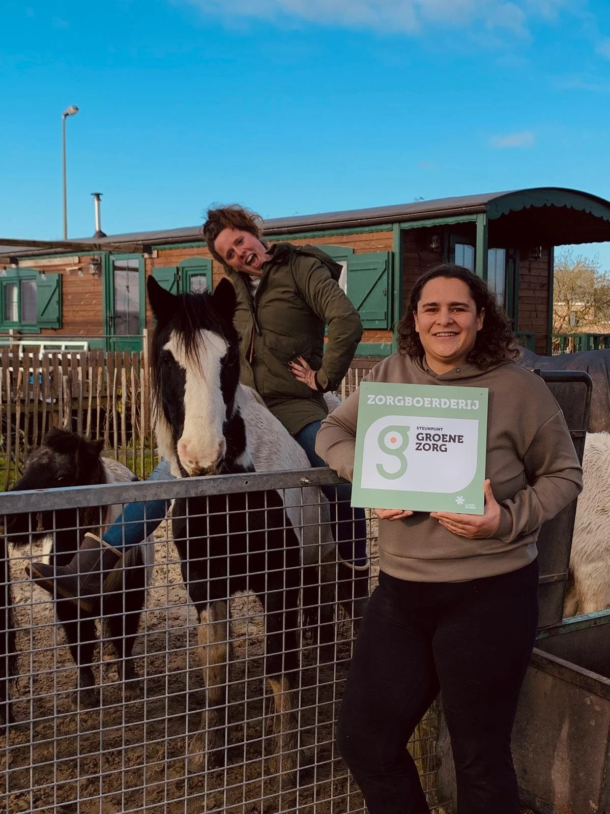 Vrouw houdt bord met tekst 'Zorgboerderij' en 'Steunpunt Groene Zorg' voor een groep dieren, met twee vrouwen op de achtergrond, een op een paard en de ander lachend.
