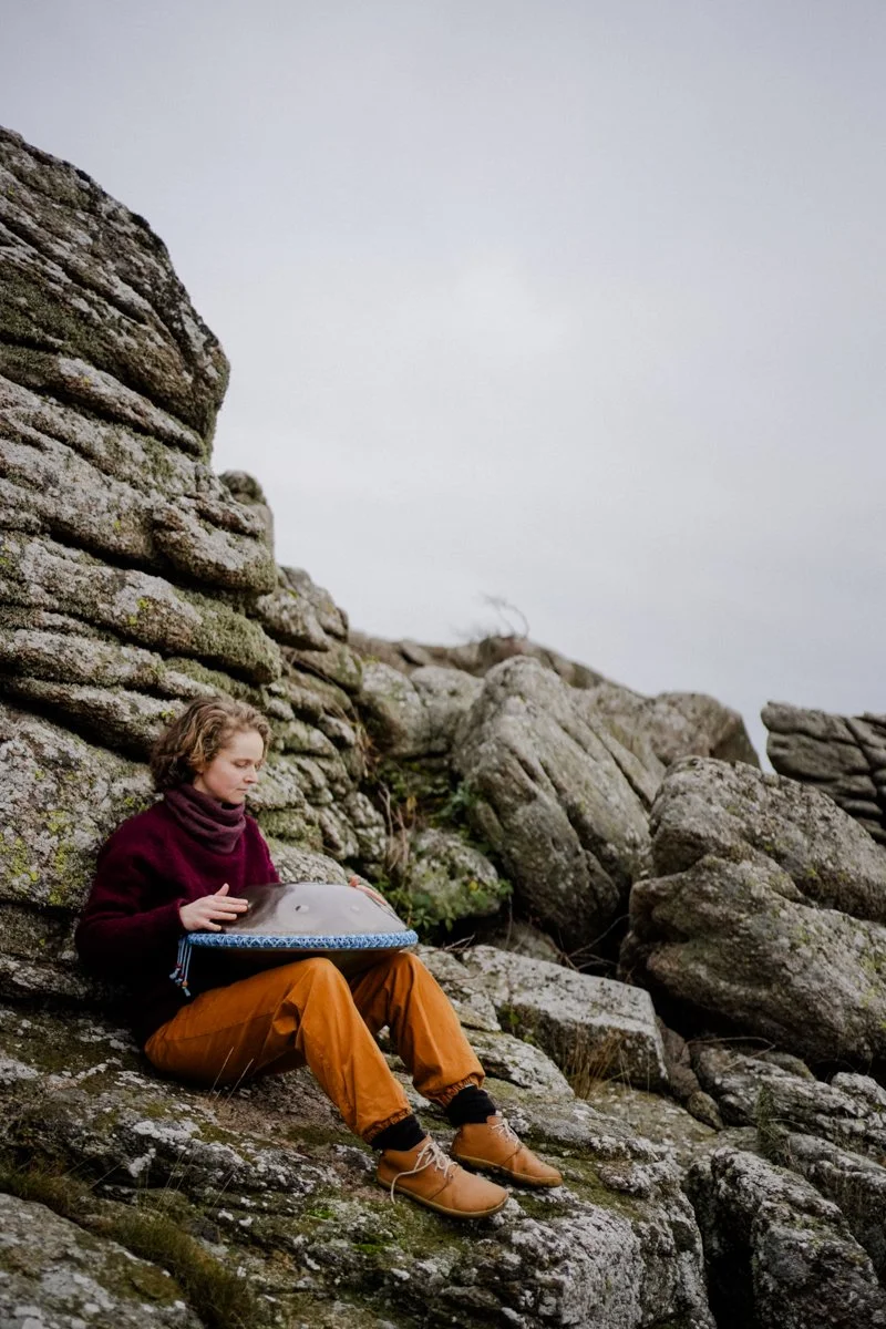 A woman is playing the handpan, leaning against a big rock.