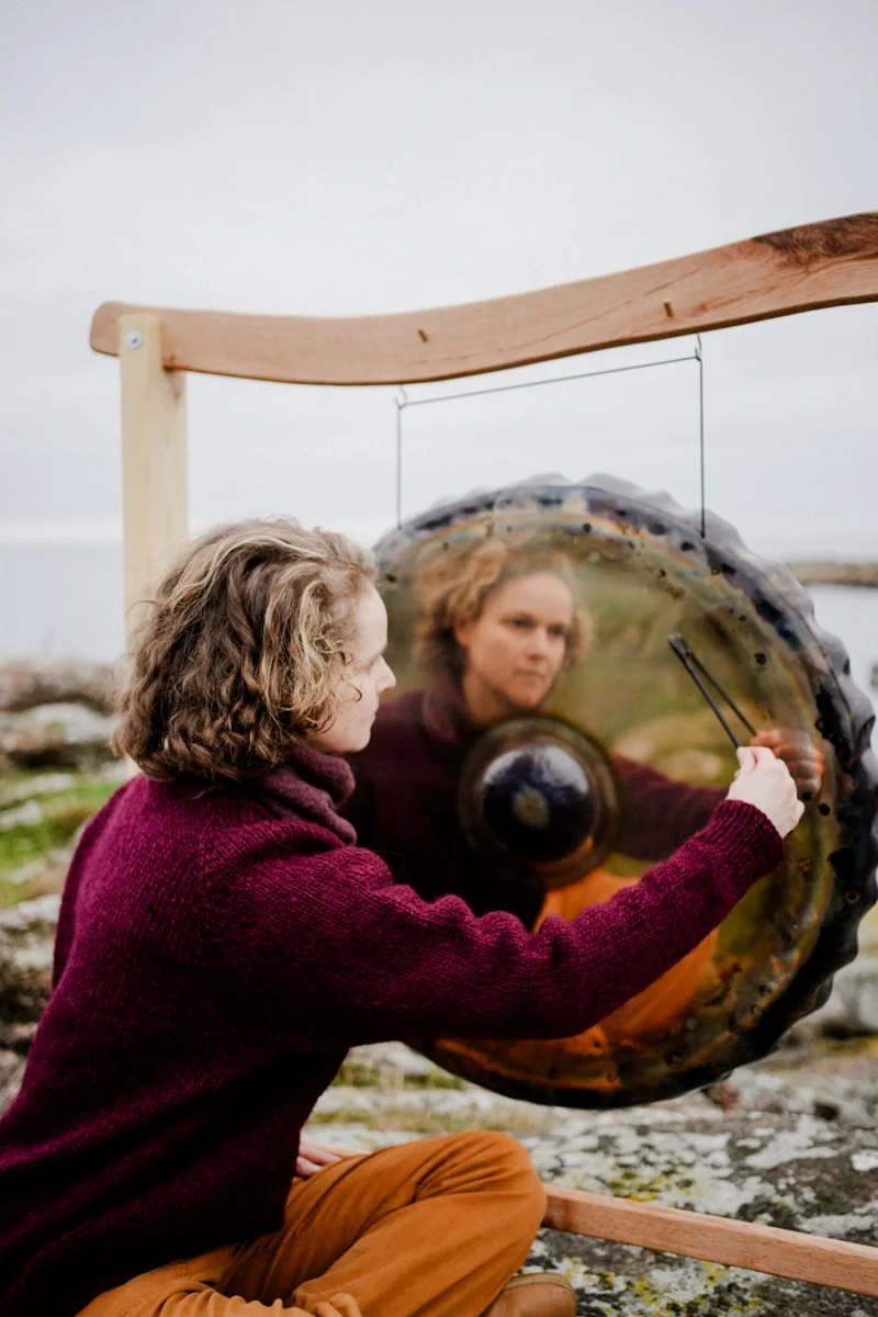 Woman playing a large circular gong outdoors, with her reflection visible in the gong's surface.
