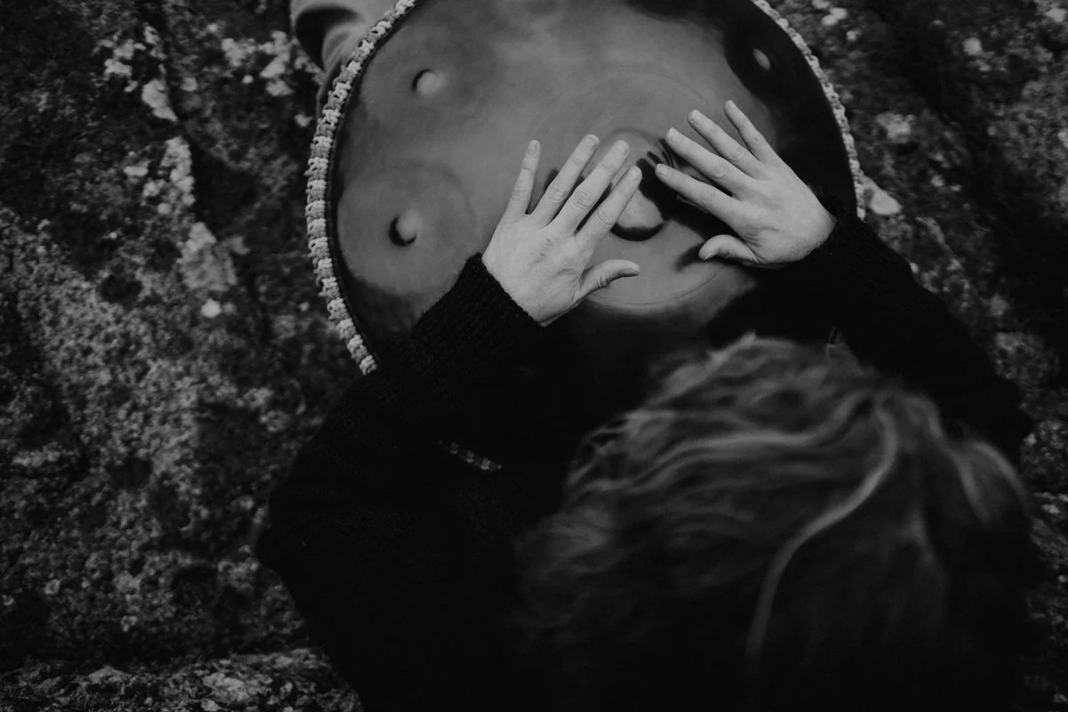 A person with light-colored hair is playing a steel drum while lying on the ground, with their hands on the drum.