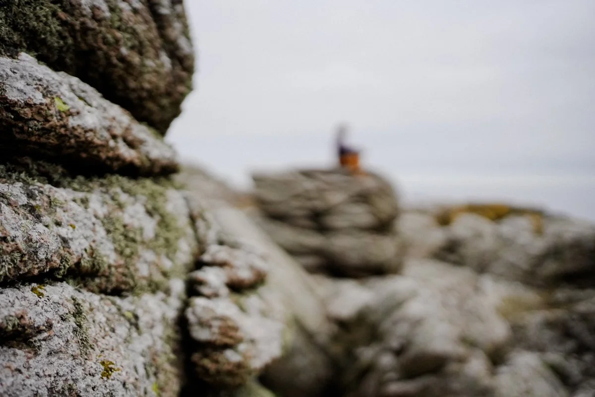 Close-up of a weathered stone wall with moss, with blurred rocks and a person sitting on rocks in the background.