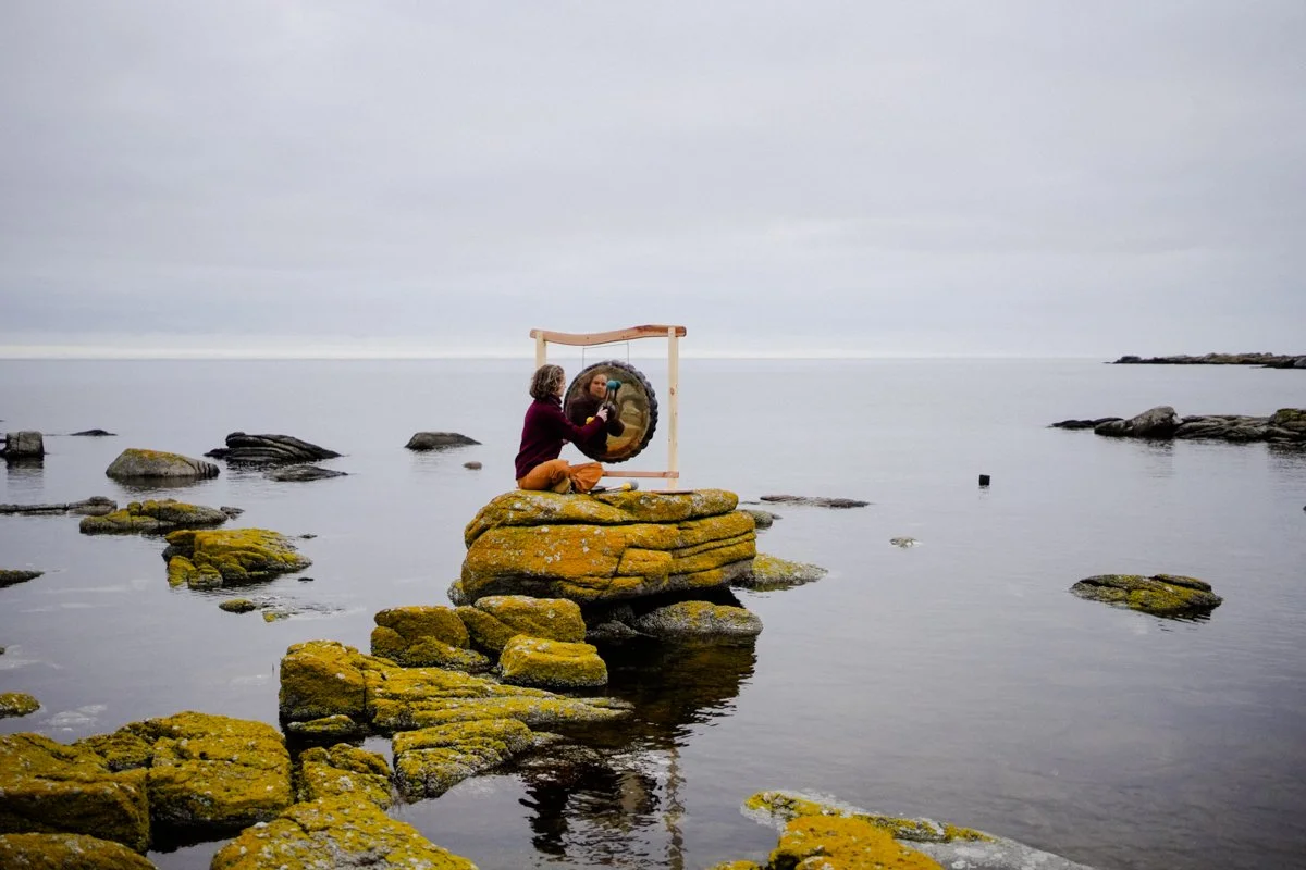 A woman is playing the Gong that is placed on a rock in the sea.