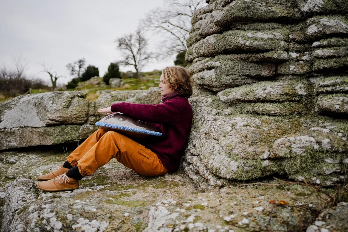 a woman is playing the handpan, leaning against a rock with grass and trees in the background