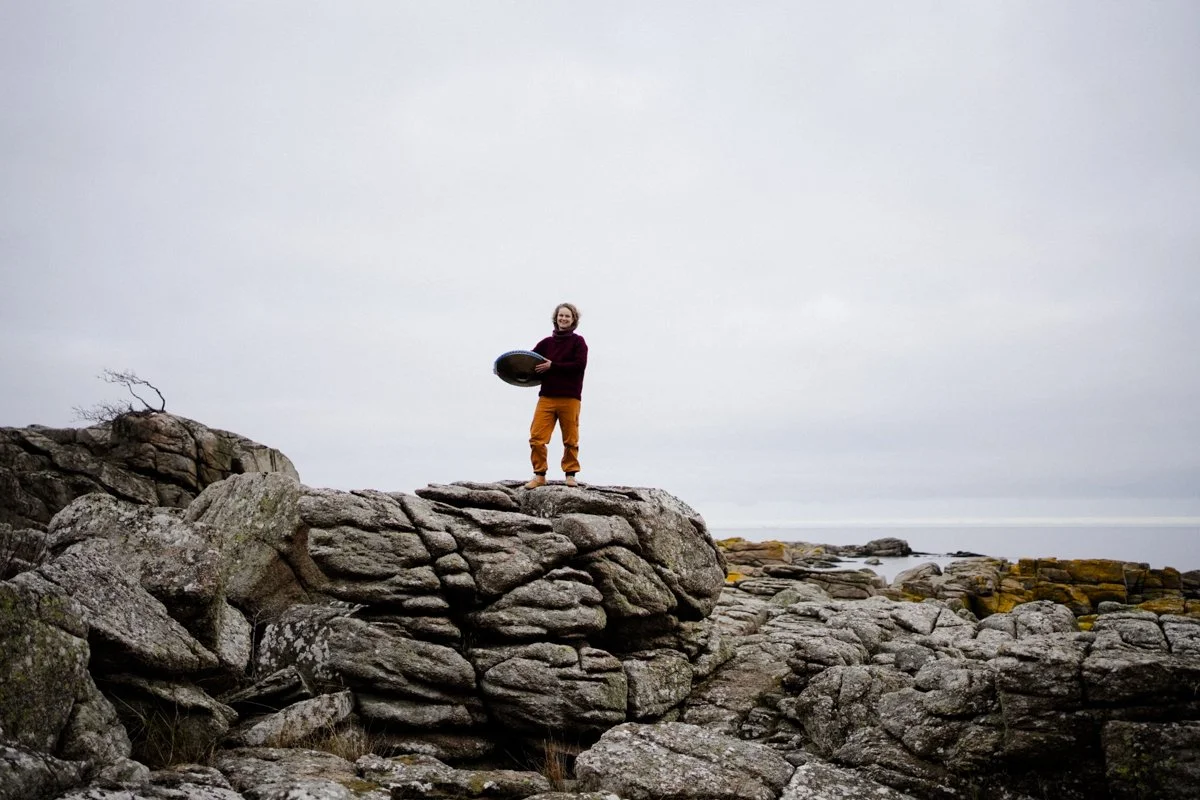 A woman is standing high on a rock, with the sea behind her. She is holding a handpan in her hands, laughing joyfully.
