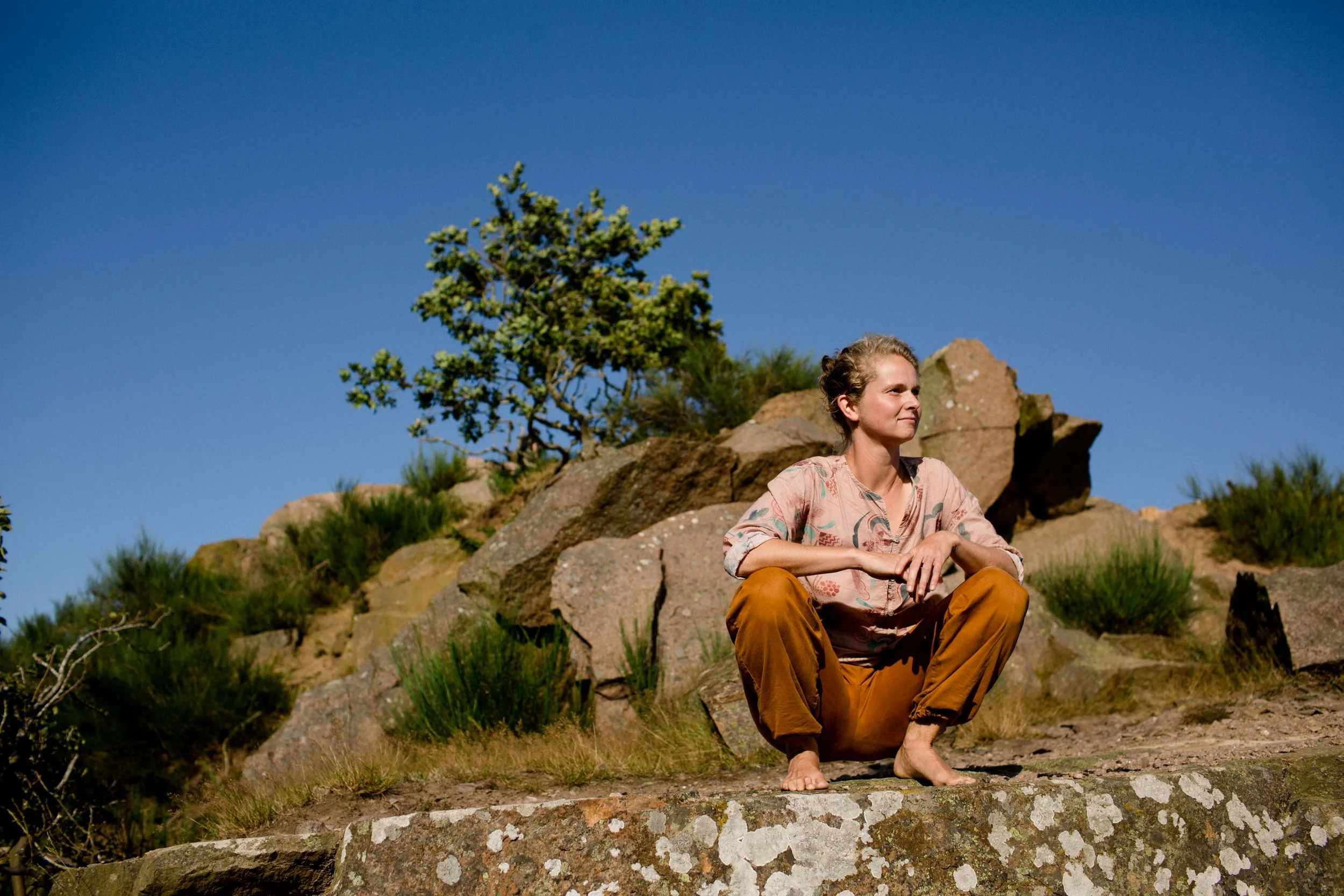 A woman sitting in squat-pose on a rock, looking out into the landscape. There are big rocks, some trees and bushes and a clear blue sky behind her.