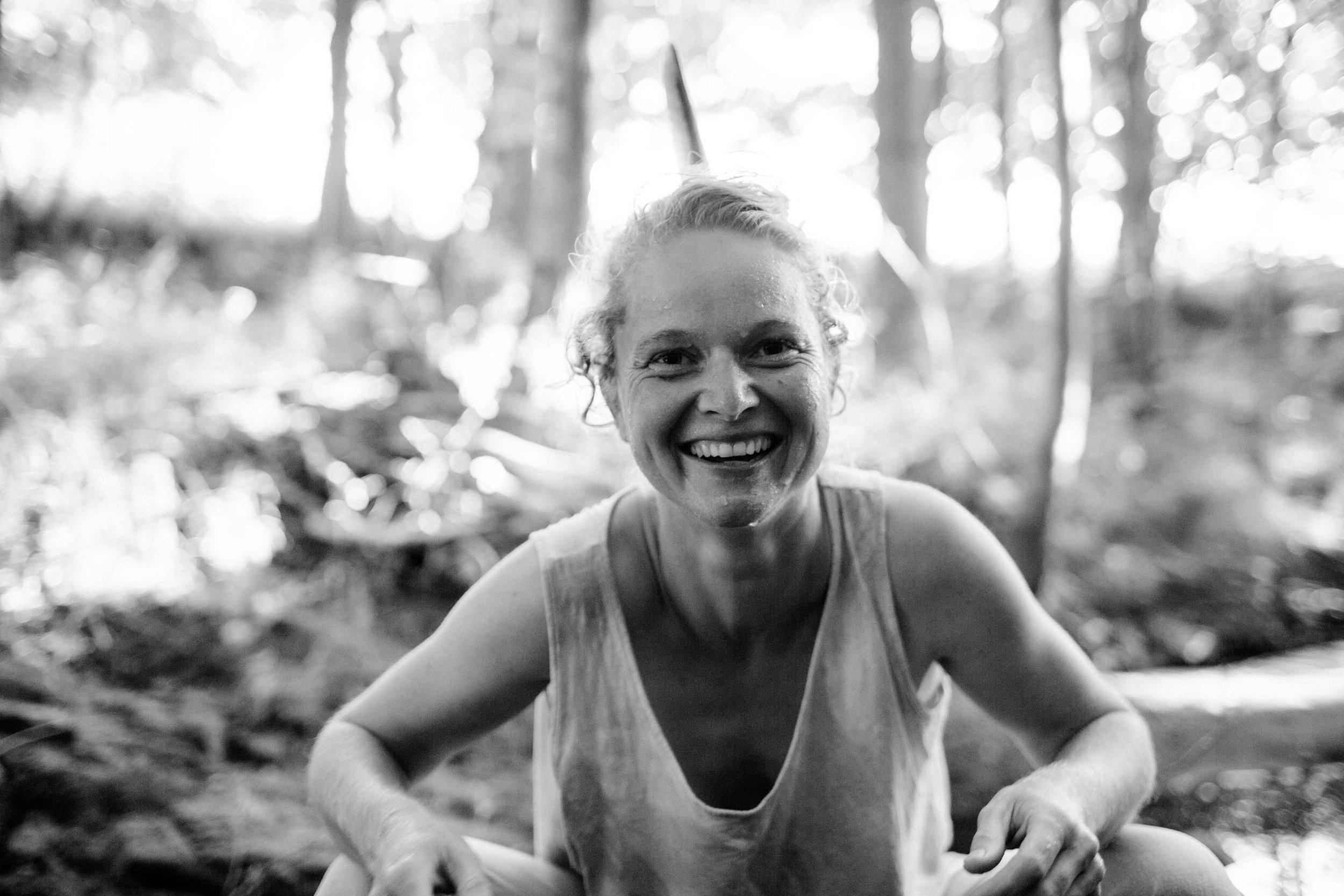 A woman is laughing wholeheartedly into the camera. She is in a forest and has drops of water on her face.