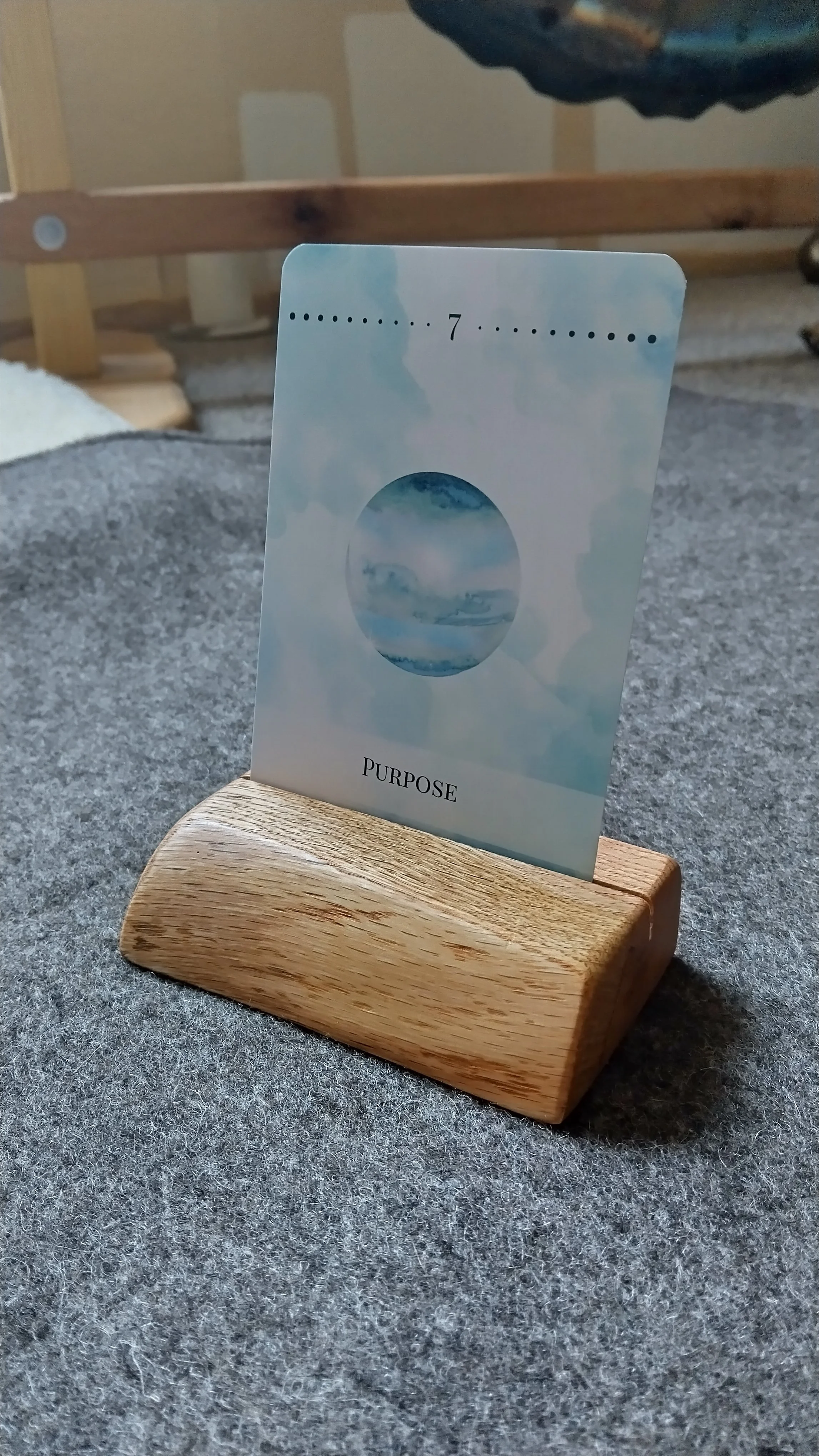 Ein Motivationskartenhalter mit einer Karte, die den Begriff 'Purpose' und die Zahl 7 zeigt, in einem Holzhalter auf einem grauen Stofftuch.