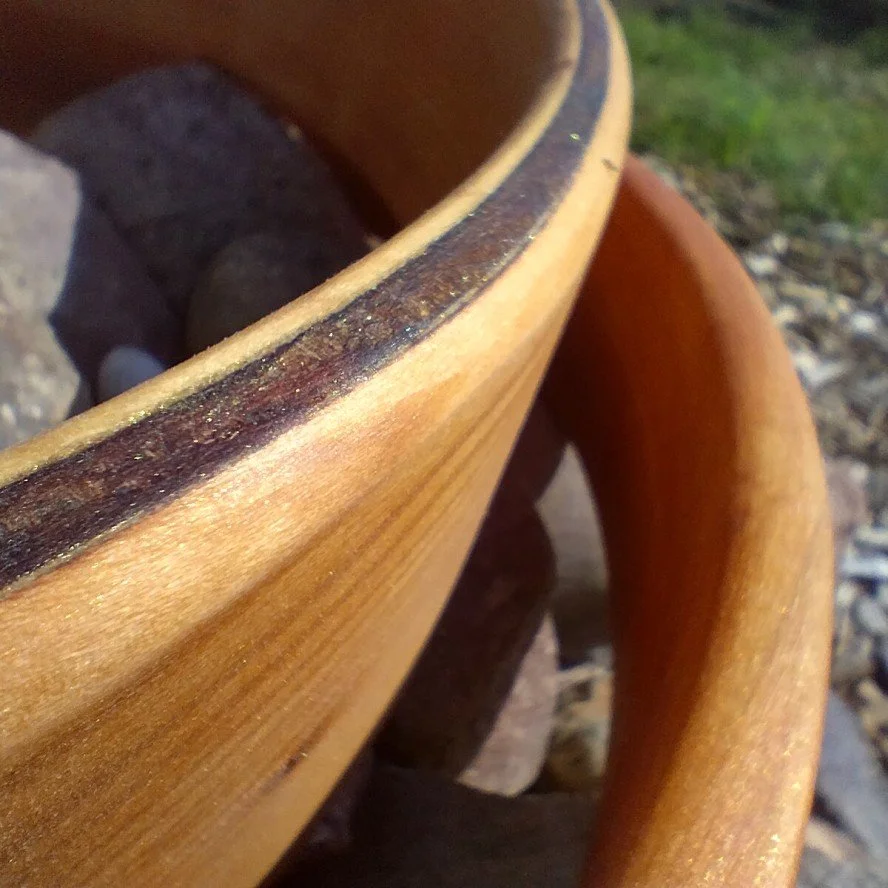Close-up of a terracotta plant pot with a wooden handle and some rocks inside the pot, outdoors on gravel with some greenery visible in the background.