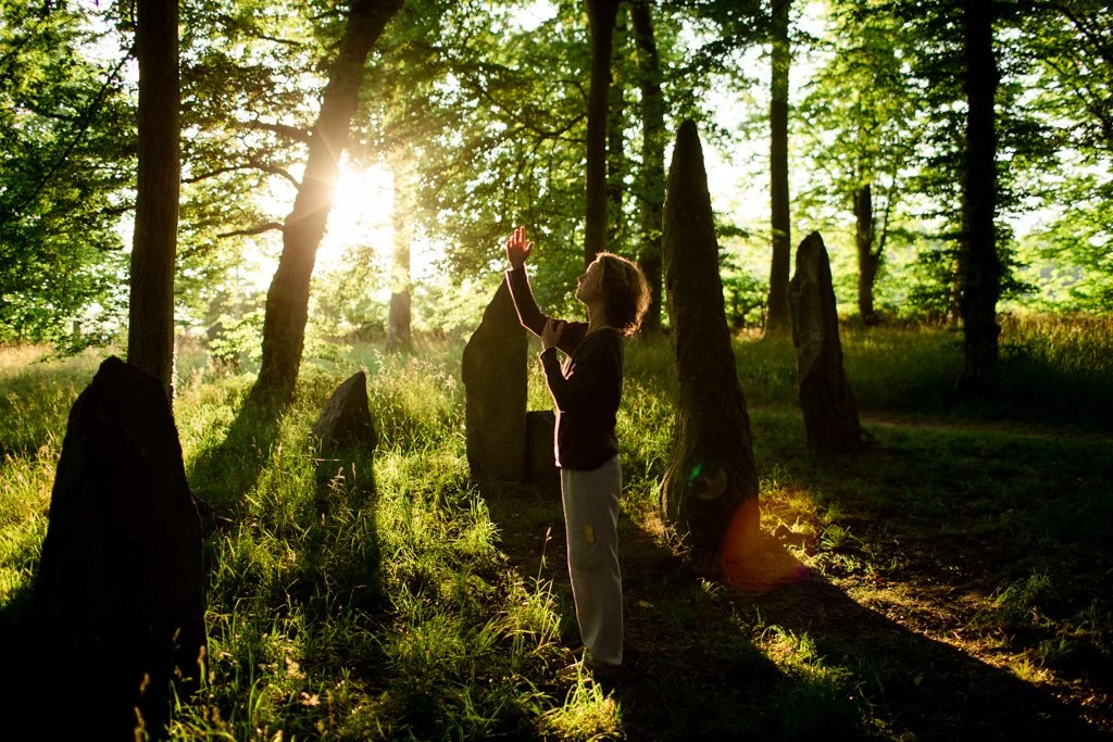 A person practicing yoga or meditation outdoors in a forest during sunset, surrounded by standing stones or large rocks.