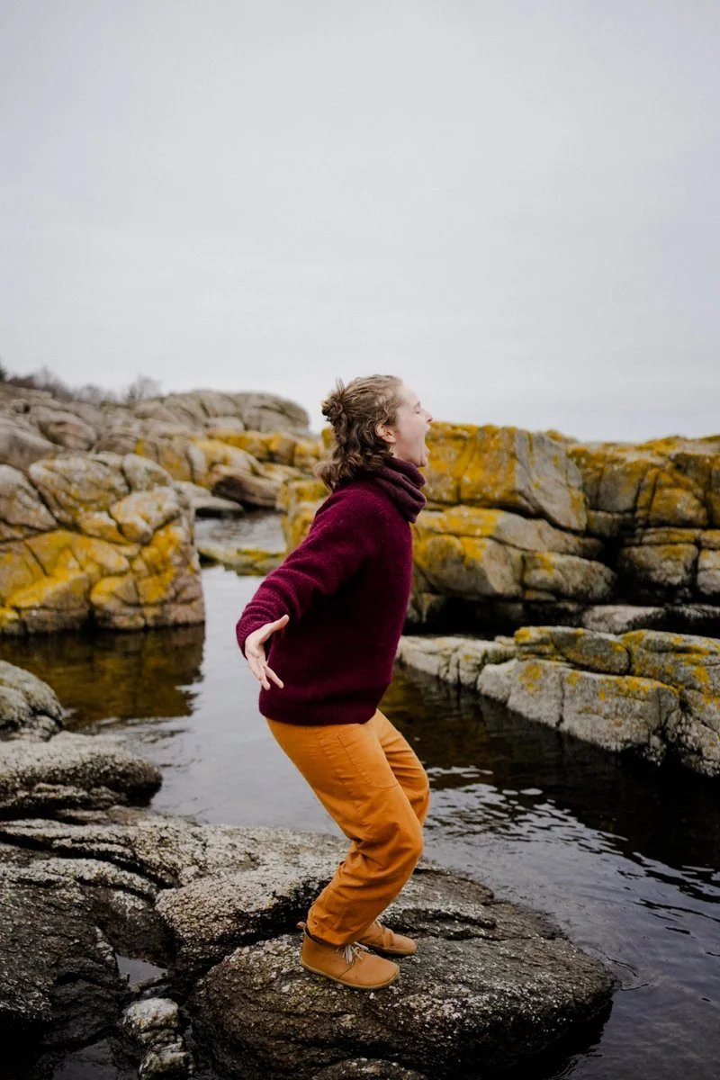 A woman standing on rocks near water, yelling or screaming with her mouth wide open, outdoors in a rocky landscape.
