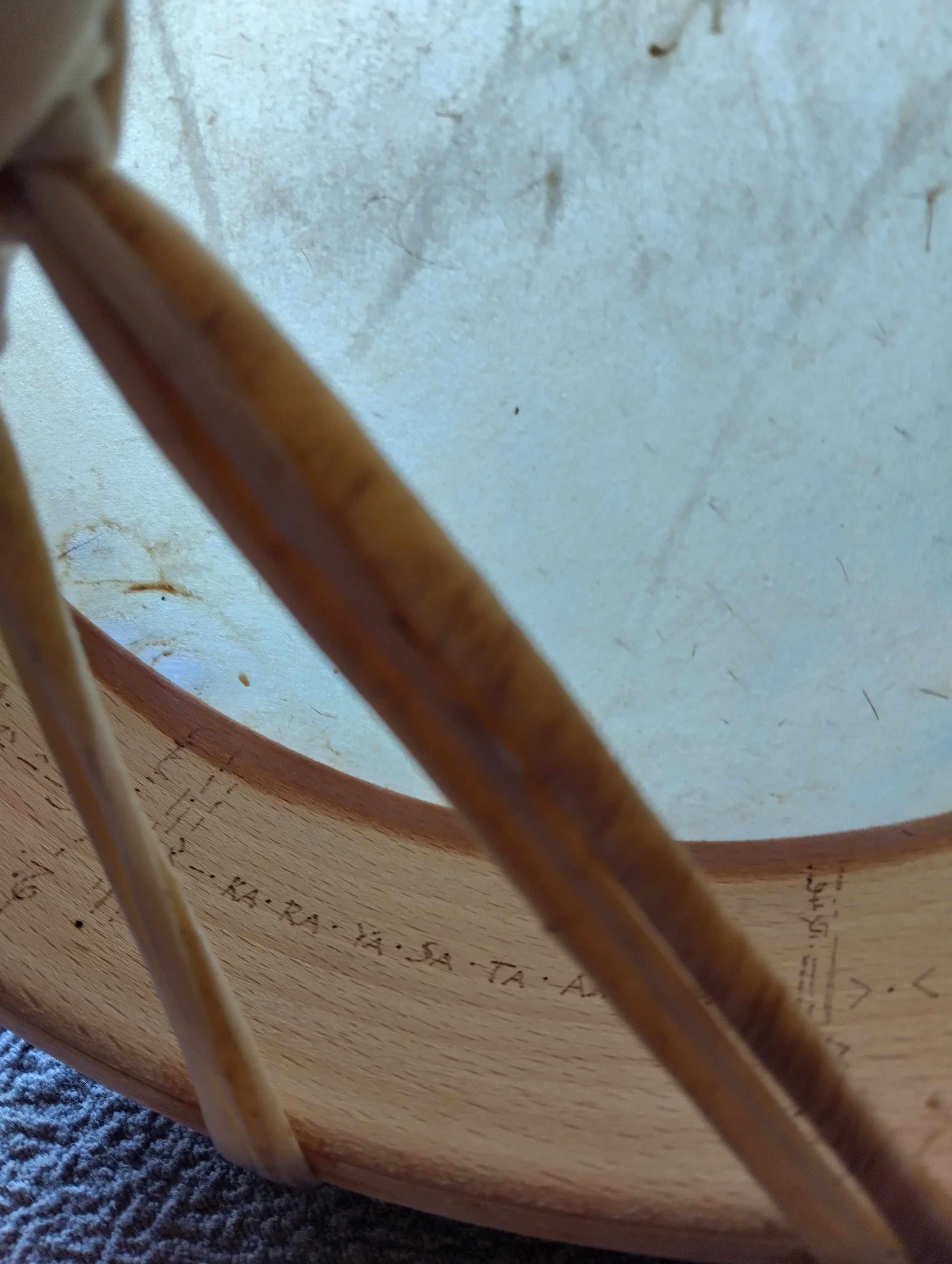 Close-up view of the underside of a wooden chair with a round base, showing the curved support rods and the printed specifications on the wood, with part of a textured grey carpet visible.