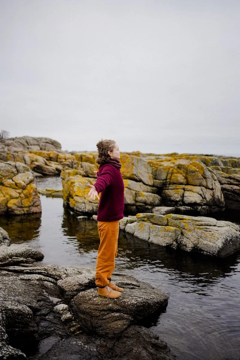 A woman is standing on a rock in the sea, arms wide open, eyes closed, inhaling deeply