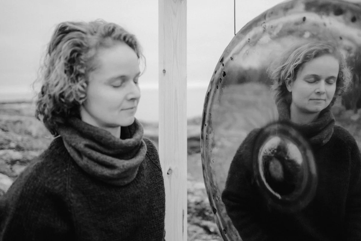 A woman is sitting outside, with closed eyes in front of a gong, the gong mirrors her face in its surface