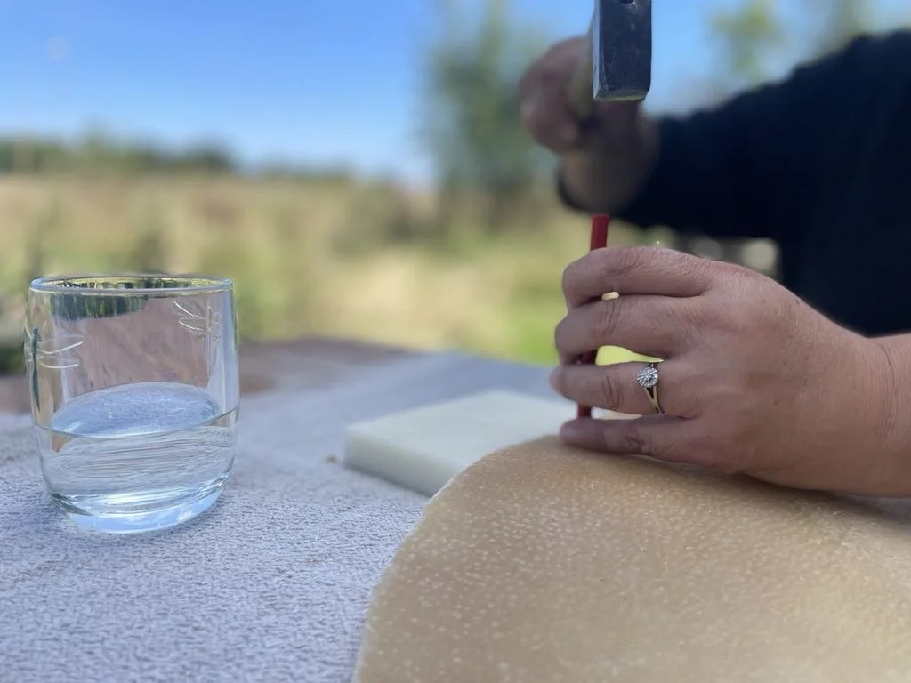 A person using a hot wire to cut a block of cheese outdoors, with a glass of water nearby.
