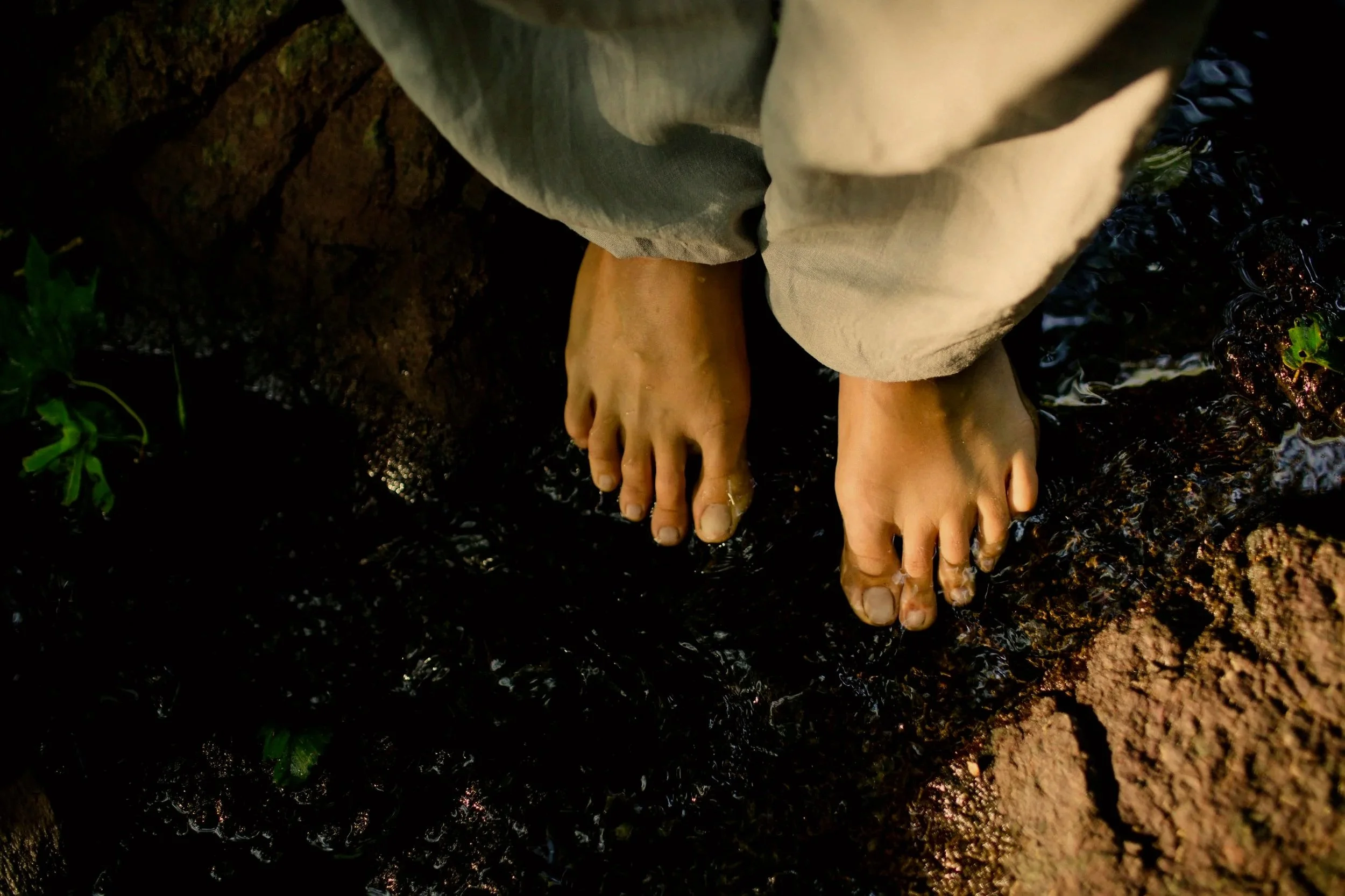 Person standing barefoot on dark wet rocks near water, wearing beige pants.