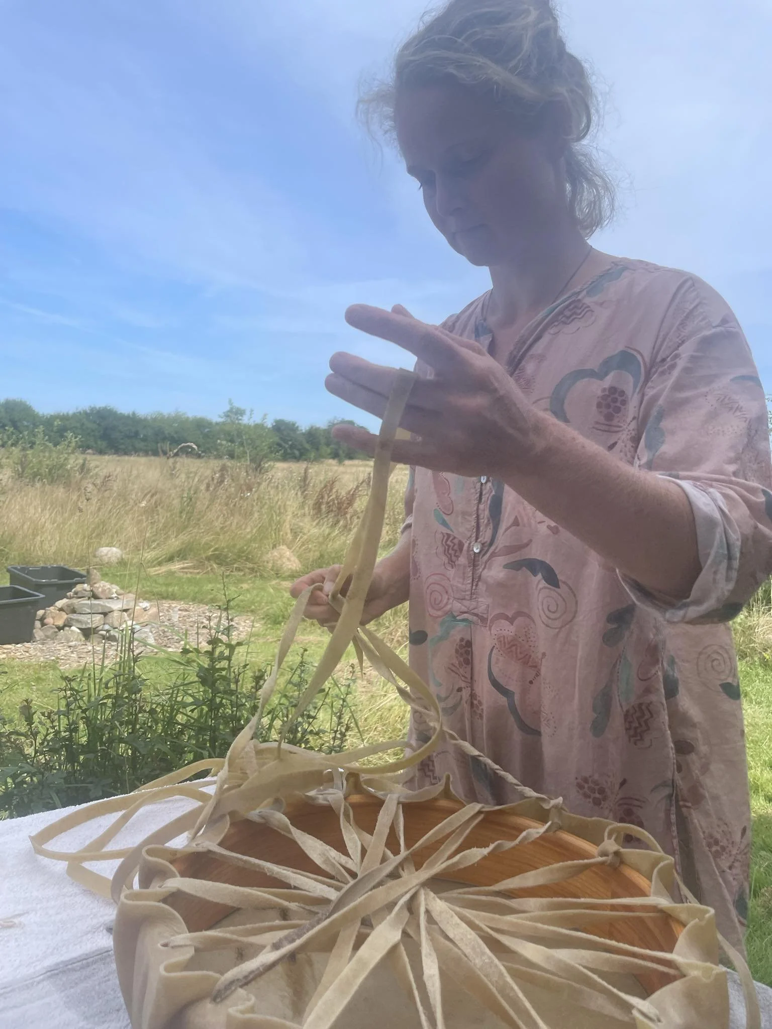 A woman in a patterned pink dress is outside under a clear blue sky, working on a craft project involving long strips of material, possibly dough or fabric, shaped in a circular or star pattern on a wooden board.