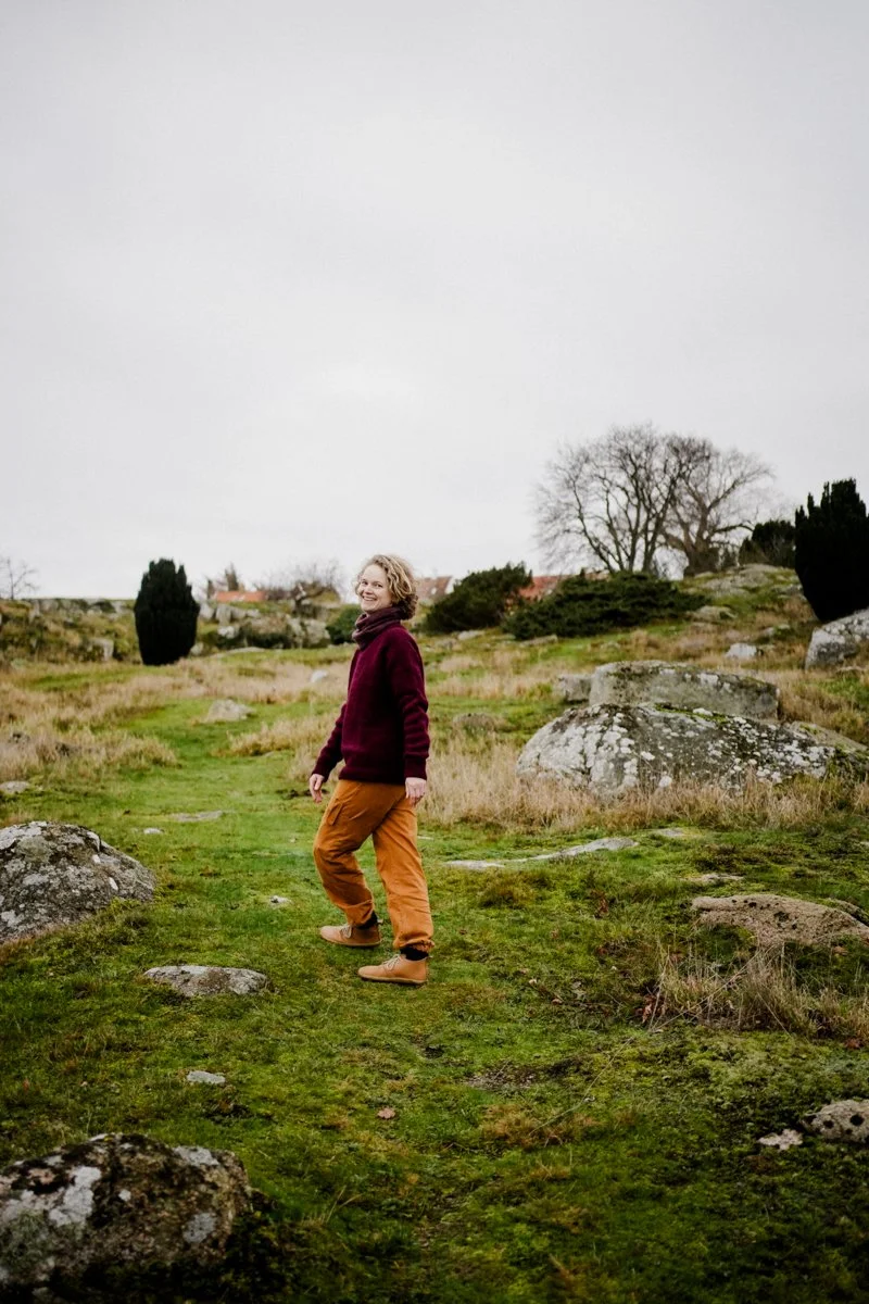 A woman is walking on a grassy path between rocks, looking back into the camera