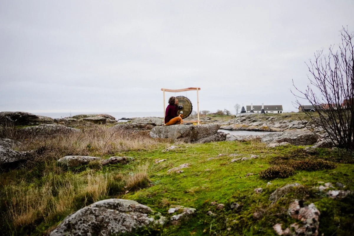 Person sitting on rocks outdoors playing a large gong with a mallet, surrounded by grassy and rocky landscape with a few houses in the distance under a gray overcast sky.