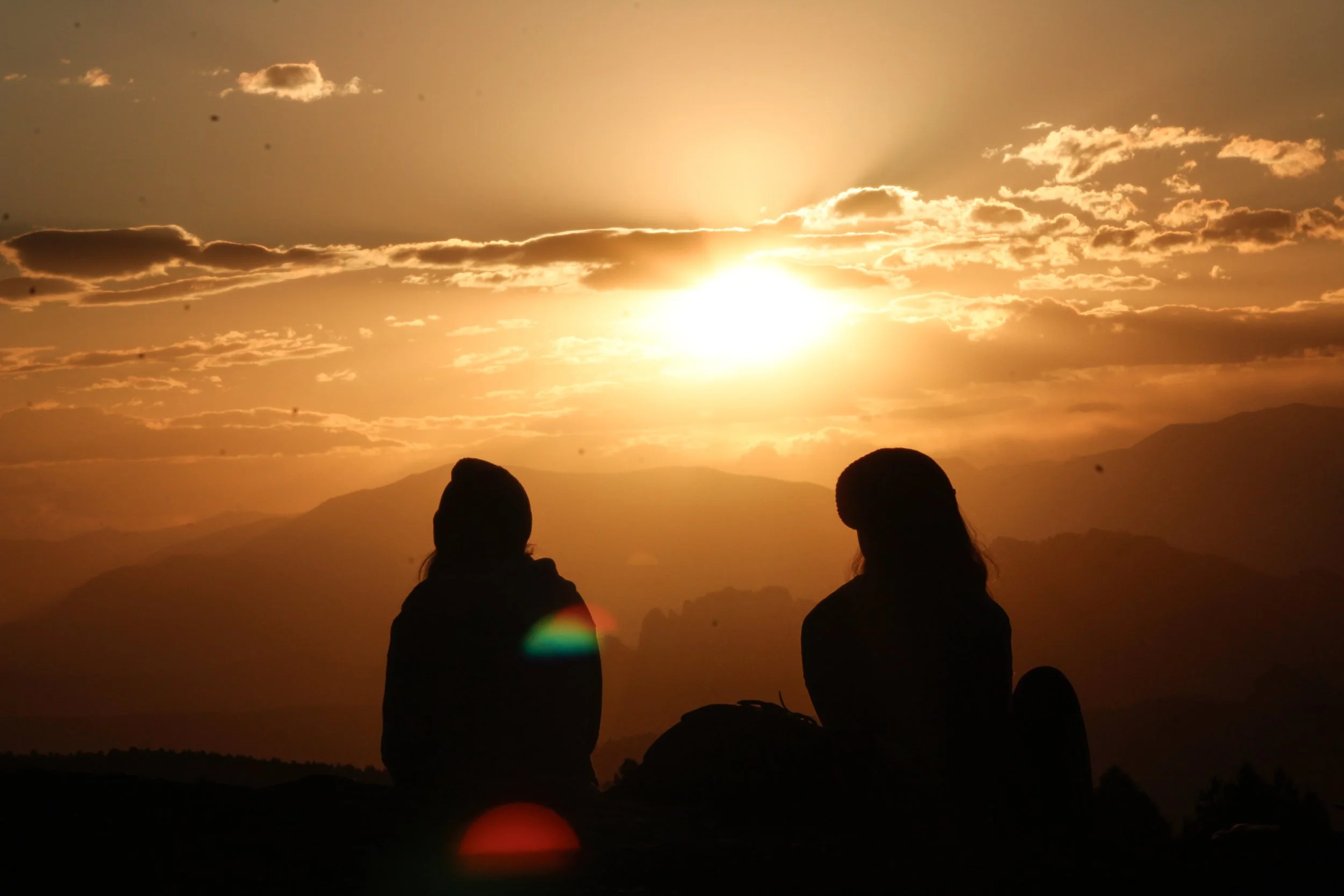 Two people sitting on a hill watching a sunset over mountains, with clouds and lens flare.