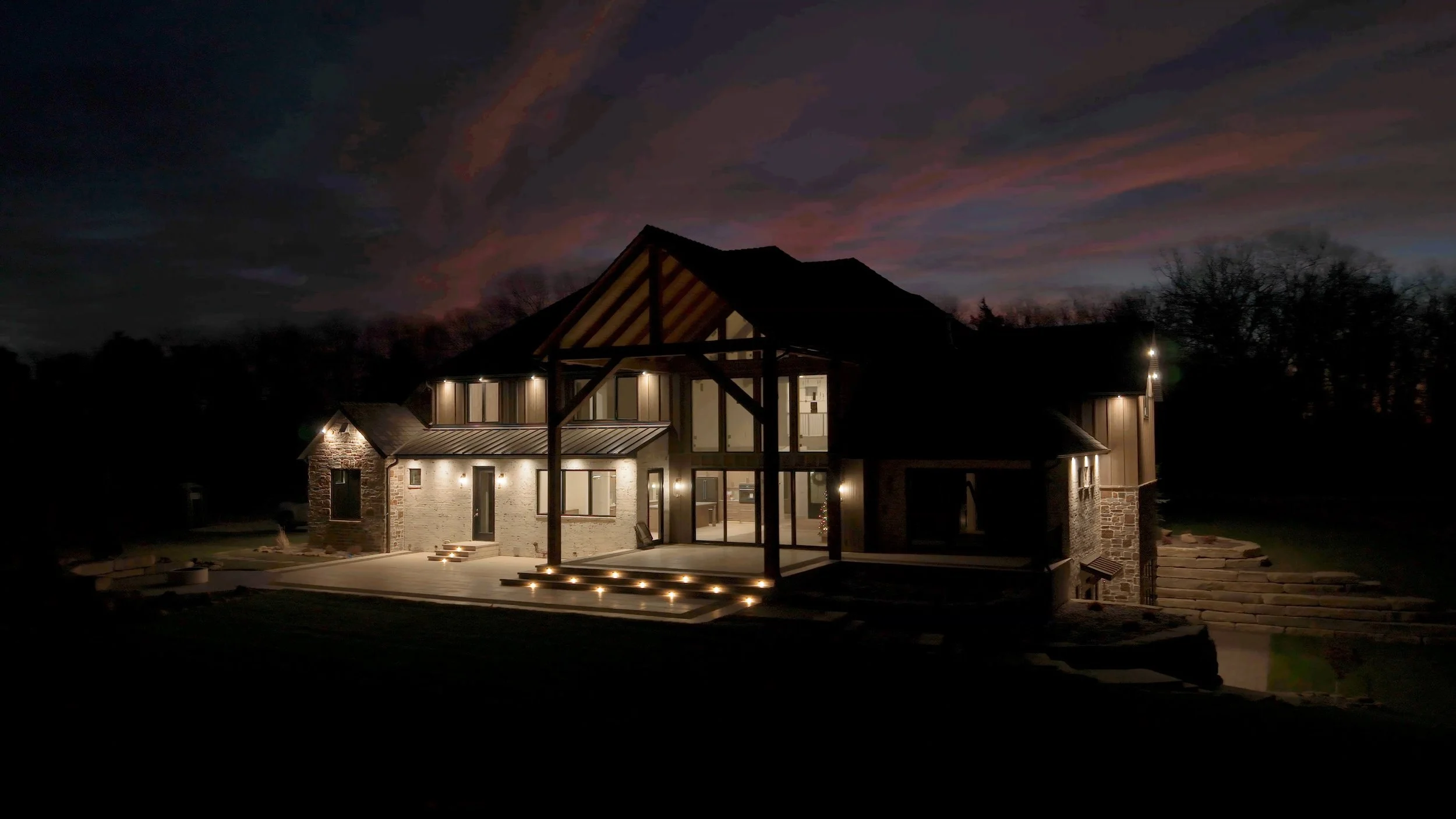 A large modern house illuminated at dusk with a colorful sky in the background. The house features stone and wood exterior, multiple windows, and outdoor lighting.