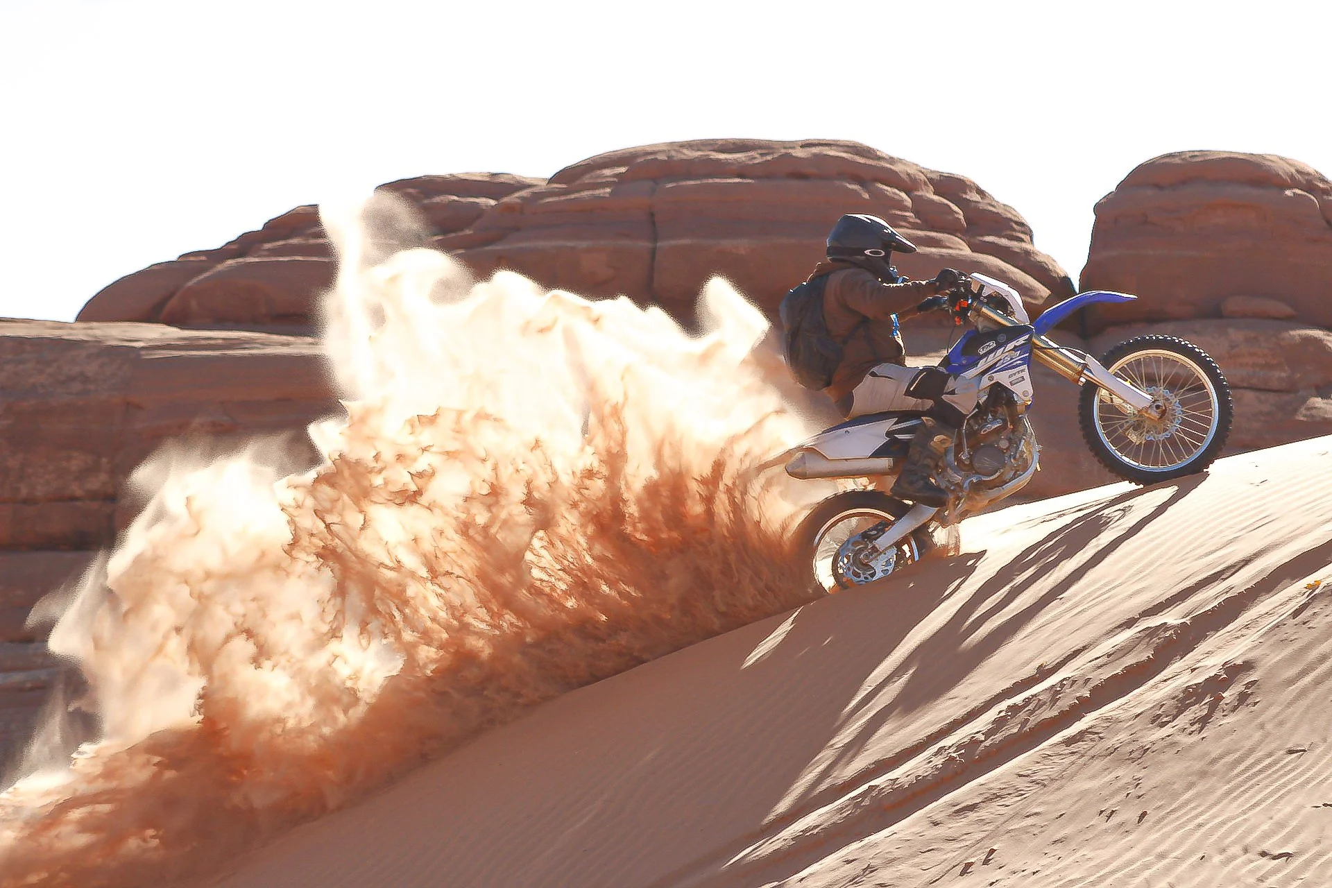 A person riding a dirt bike on a sandy dune, kicking up a spray of sand and dust, with rock formations in the background.