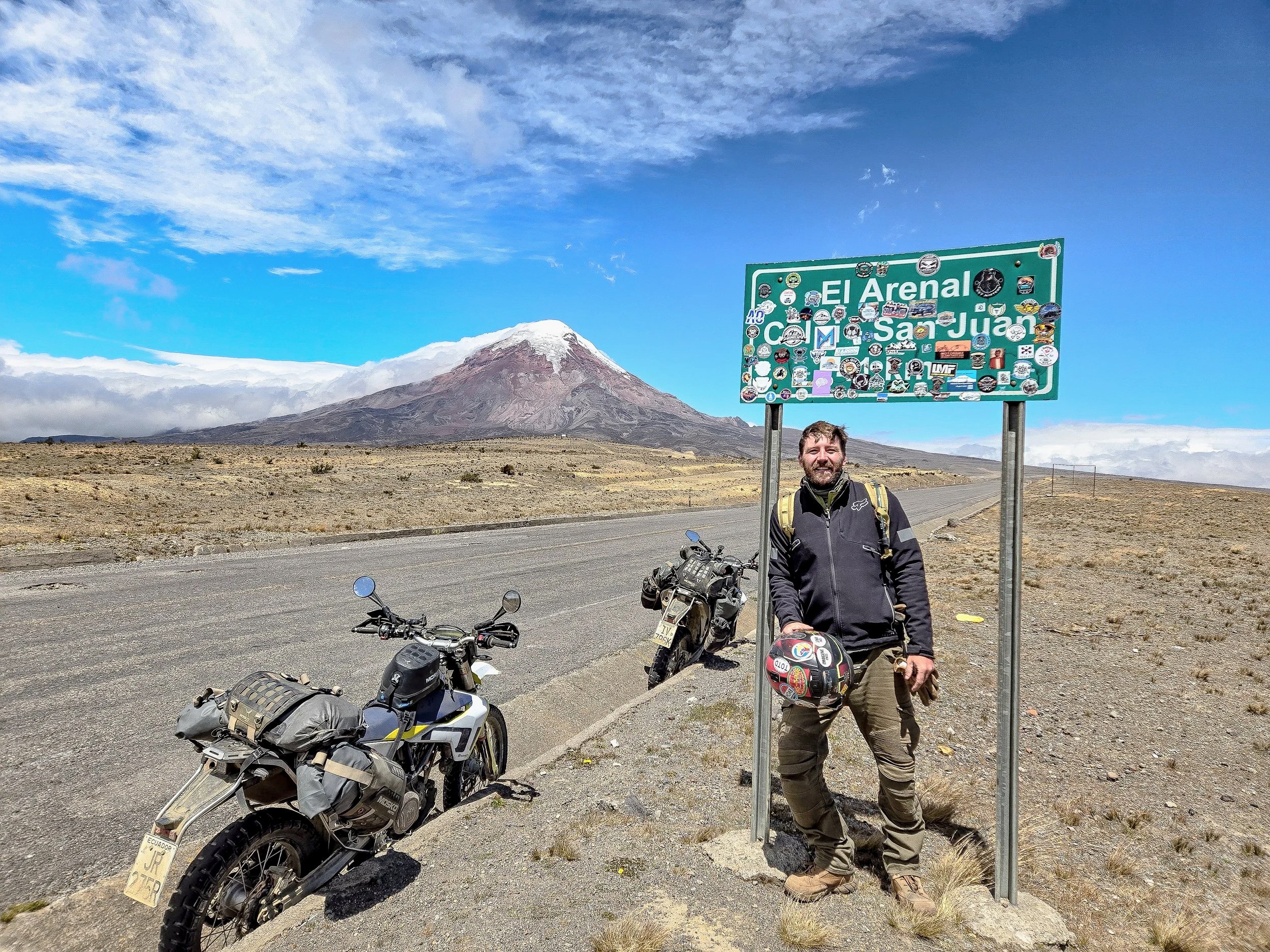 A man standing next to a signpost with numerous stickers, holding a helmet, in a desert landscape with Mount Santiago in the background.