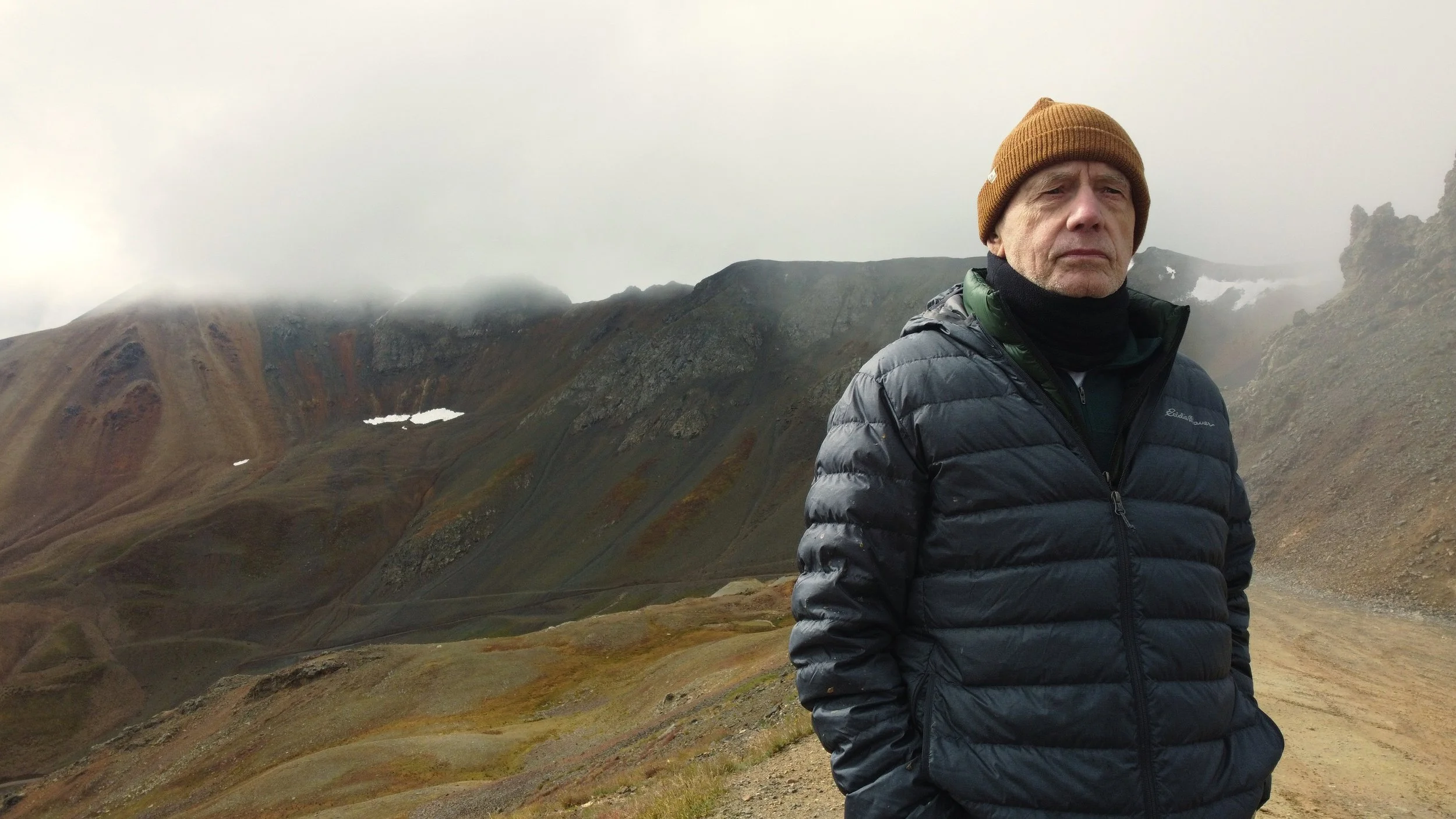 A man in a brown beanie and black puffer jacket standing on a mountain trail with rugged, dried landscape and cloudy sky in the background.