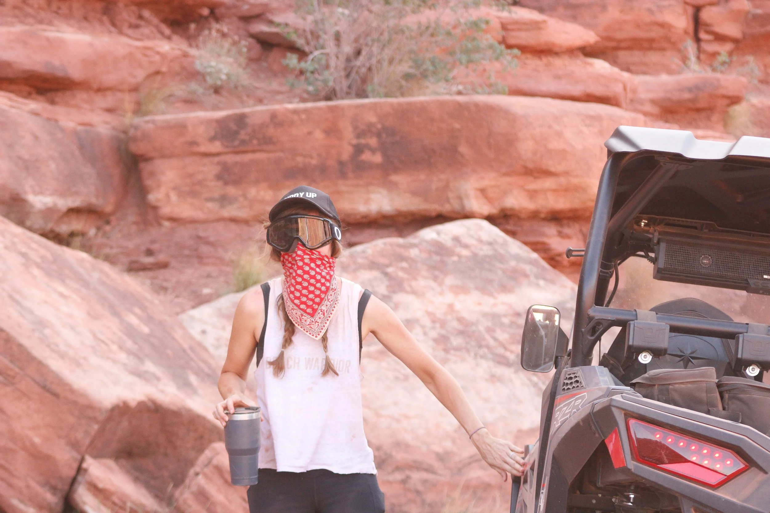 Person wearing a black cap, goggles, a red bandana covering the face, white tank top, and black pants, holding a tumbler in one hand and touching a black all-terrain vehicle in a rocky desert environment.