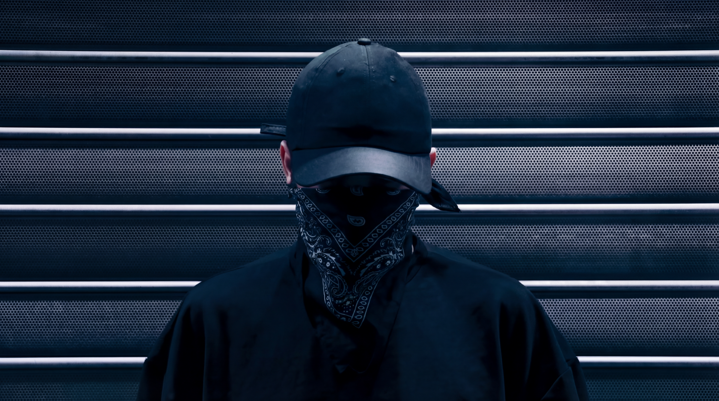 Person wearing a black baseball cap, black bandana covering face, and black jacket standing in front of a metallic background with horizontal slats.
