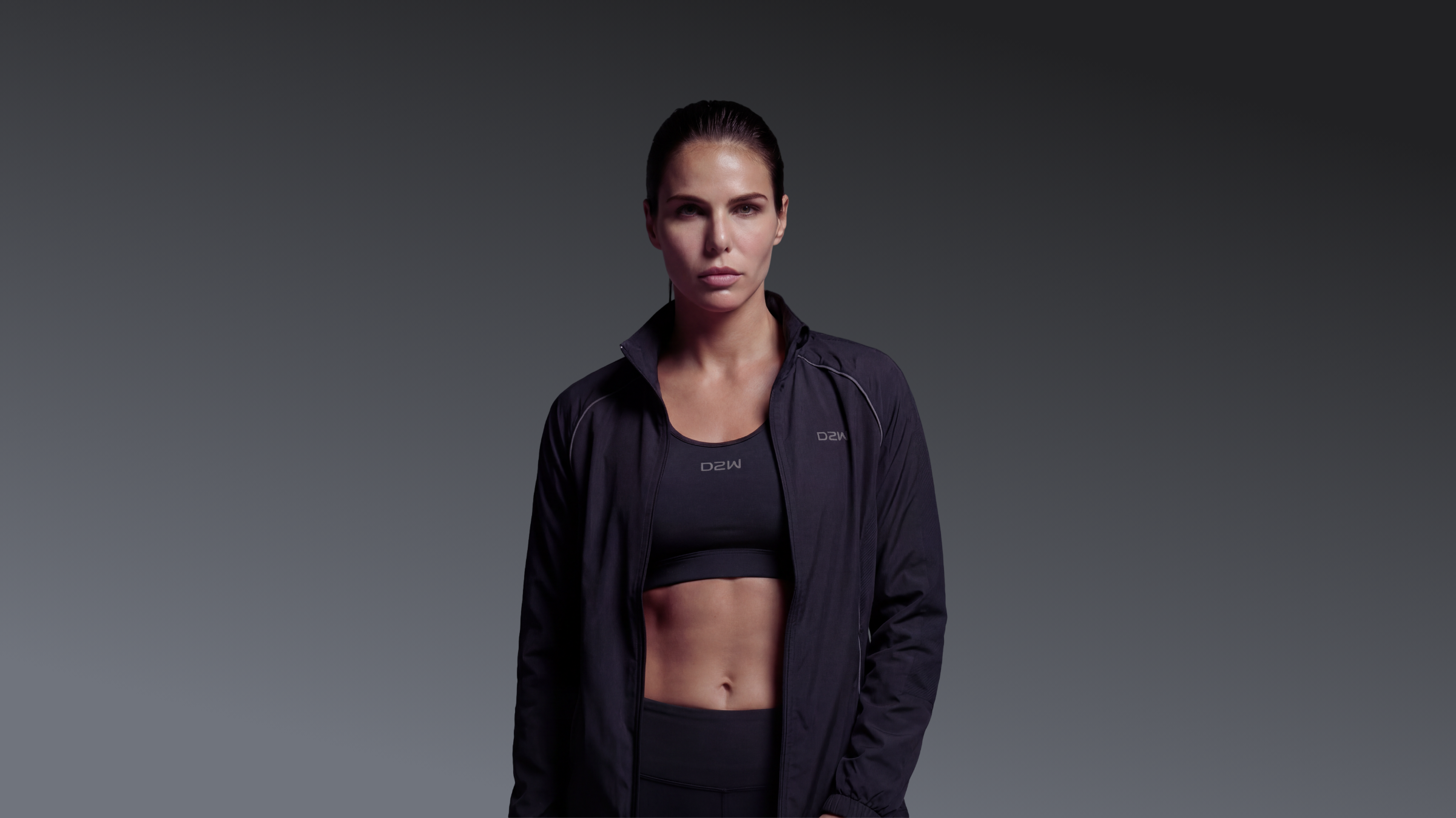 A fit woman wearing a black athletic jacket and sports bra, standing against a gray gradient background.