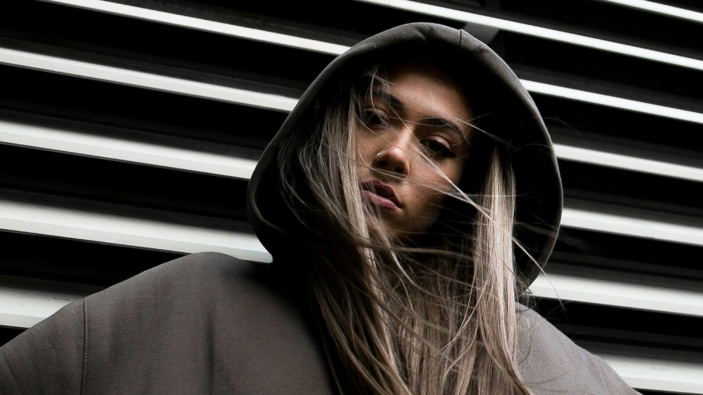 A woman with long hair wearing a gray hoodie, looking at the camera with wind-blown hair, standing in front of black and white striped background.