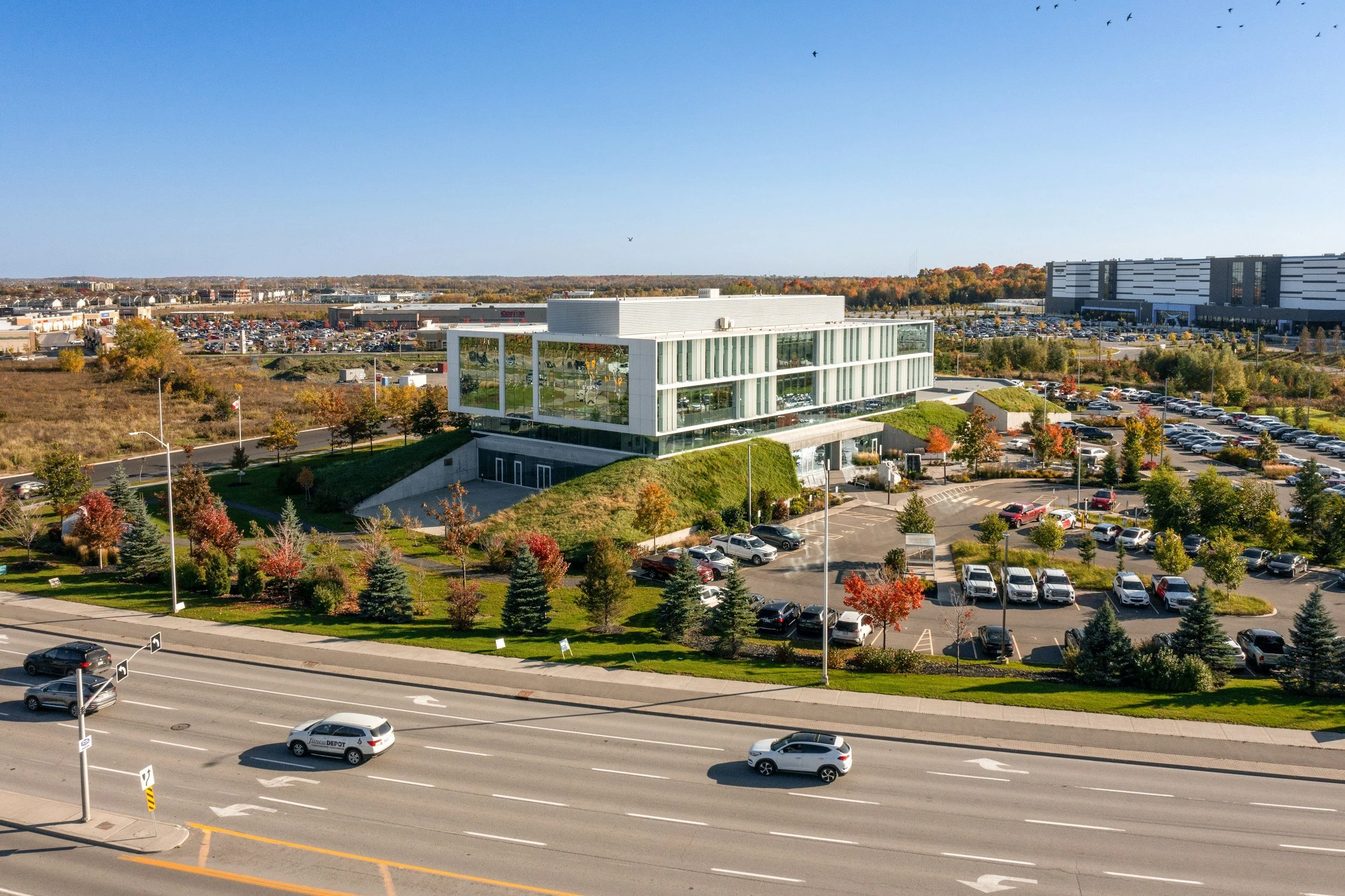 A modern, glass-fronted building with landscaped grounds and parking lot, situated near a multi-lane road with cars, in a suburban area on a clear, sunny day.