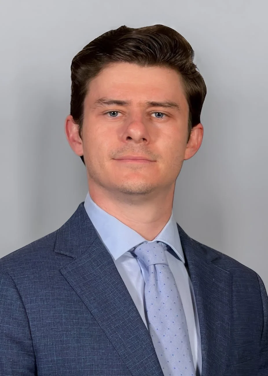 A young man with brown hair in a suit and tie, looking at the camera with a neutral expression.