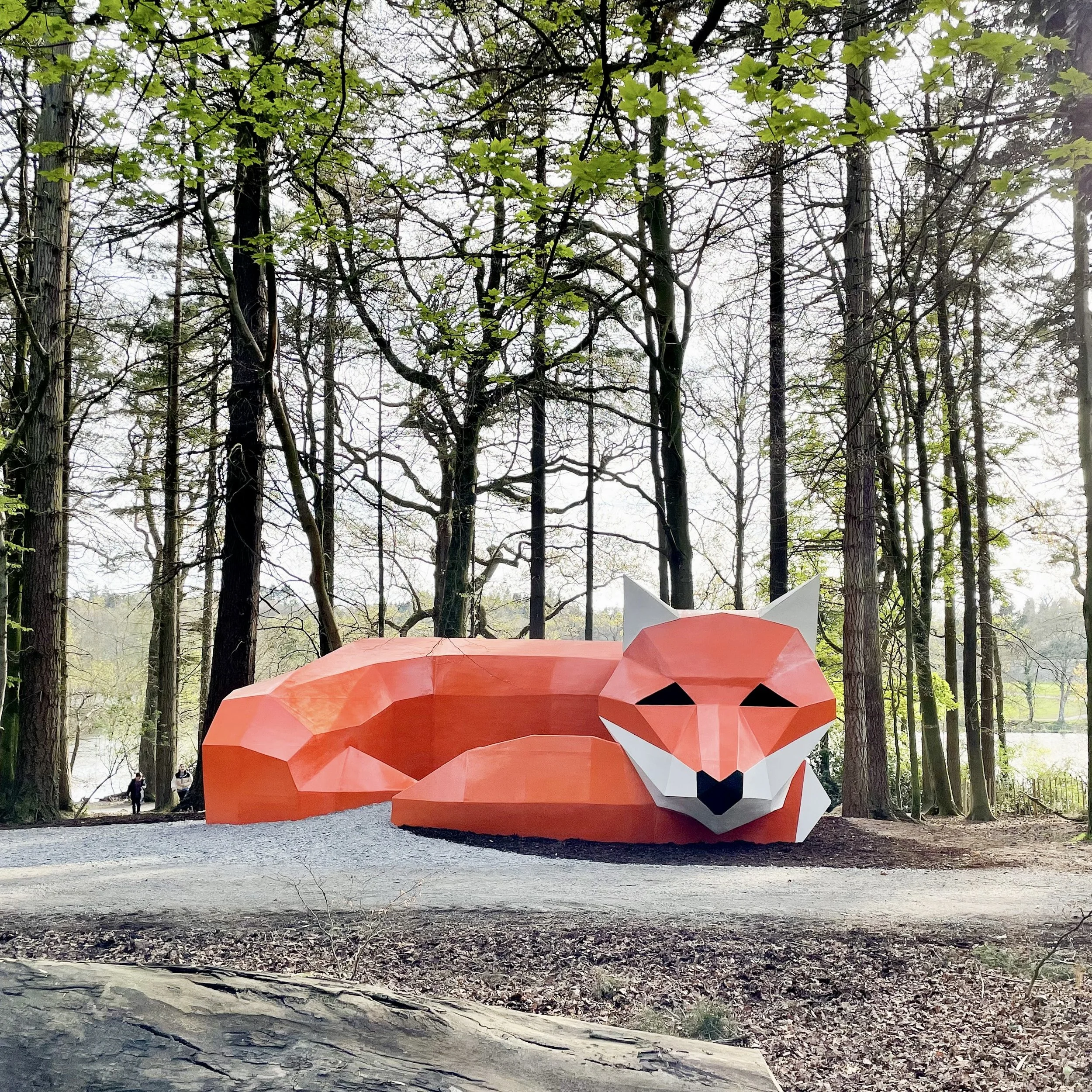 Large, geometric, red-orange fox sculpture resting on the ground in a wooded park with leafless trees and a few green leaves.